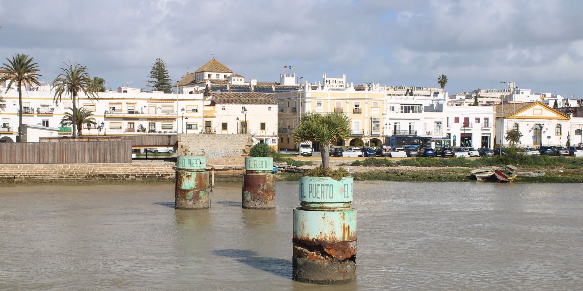 The white and ochre waterfront of El Puerto de Santa María across the Guadalete, with three rusted pillars painted El Puerto standing in the river
