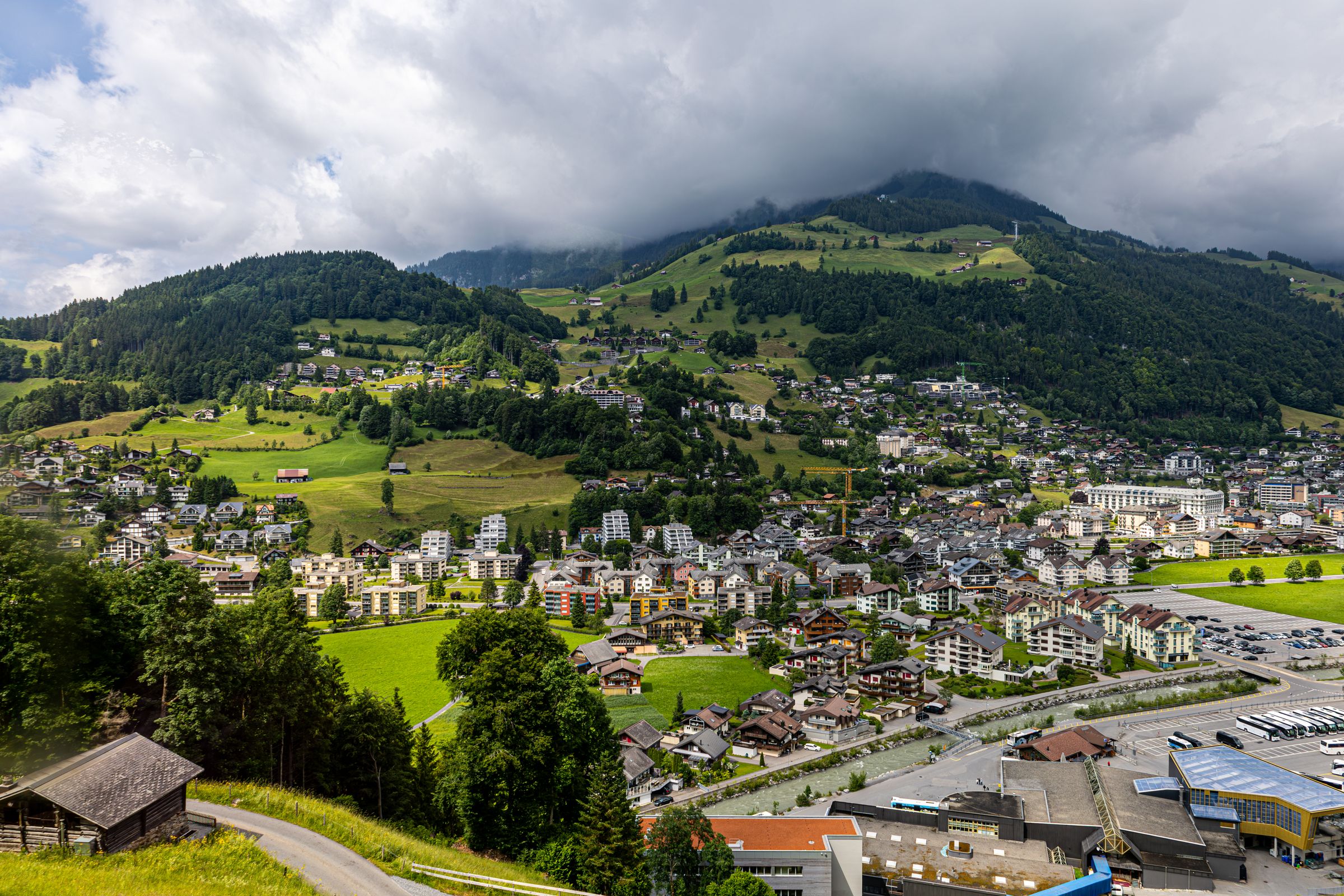 Engelberg seen from the hillside above, chalets and apartment blocks across the green valley floor under forested slopes and low cloud