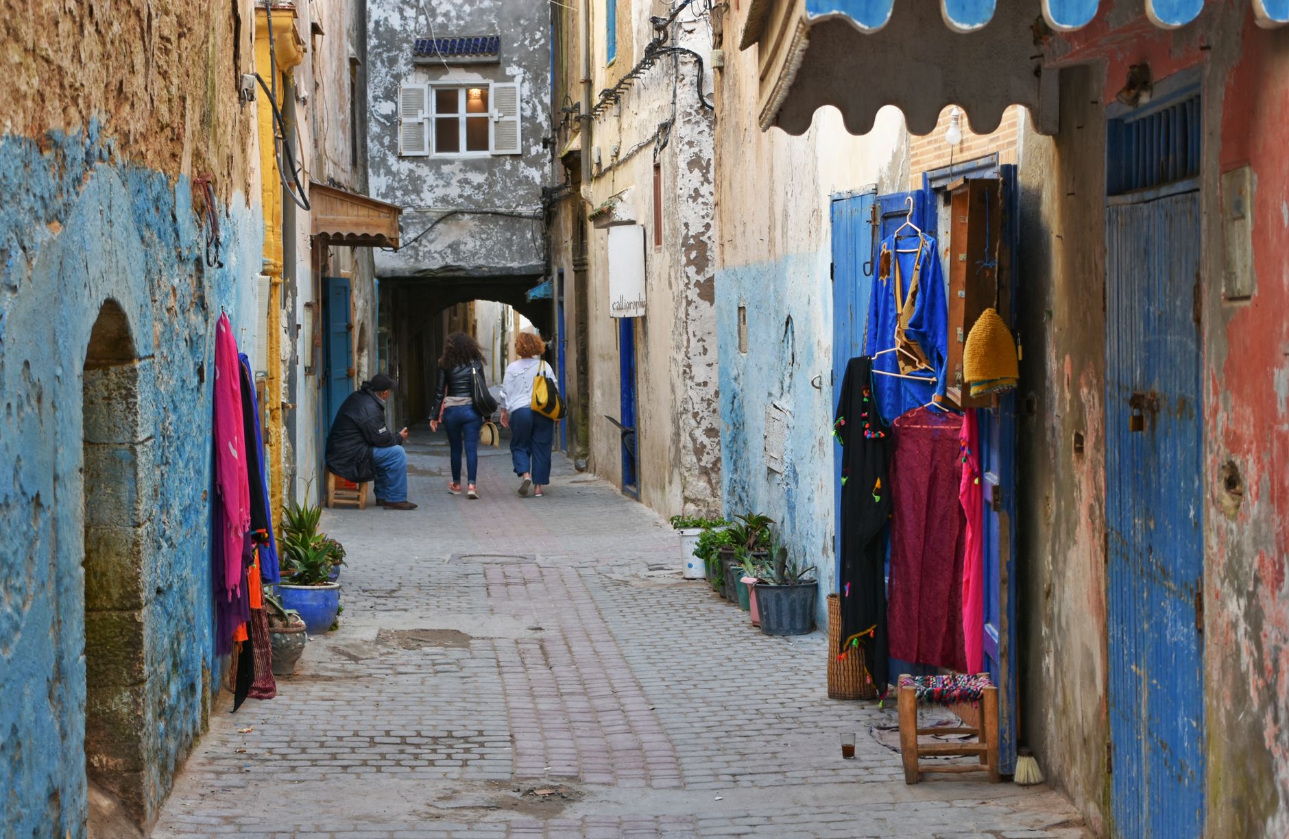 A paved lane in the Essaouira medina with blue doors, white walls and rugs and clothes hung outside a shop