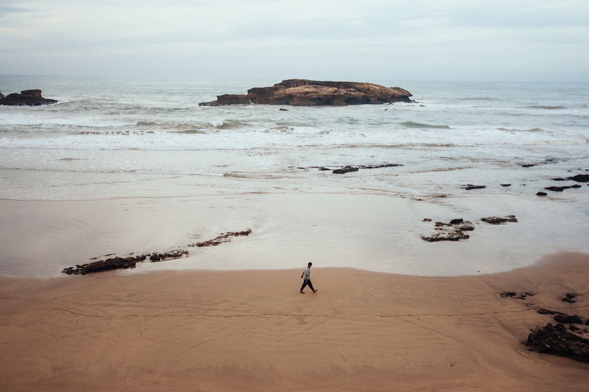 A lone walker on the wide wet sand of Essaouira beach at low water, with rocks offshore