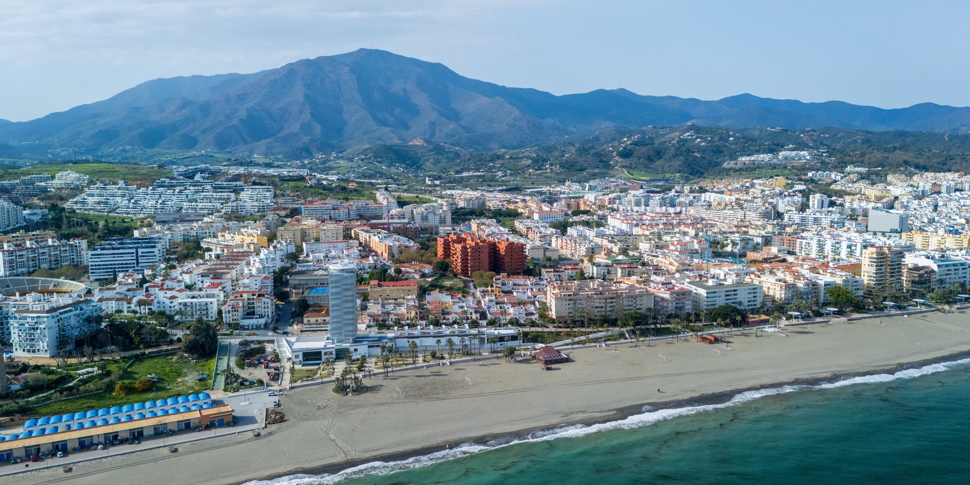 Estepona's seafront and white apartment blocks from the air, with the bullring on the left, a tall tower on the promenade and the Sierra Bermeja behind