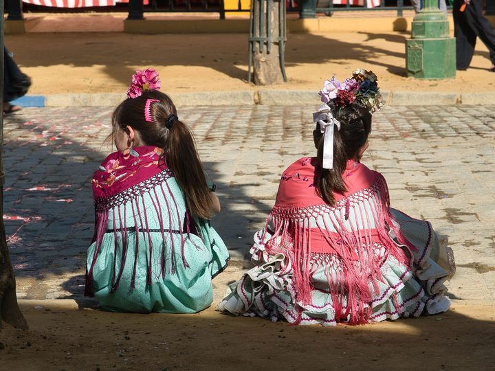 Two girls in flamenco dresses and fringed shawls sitting at the edge of a sandy street at the Seville fair