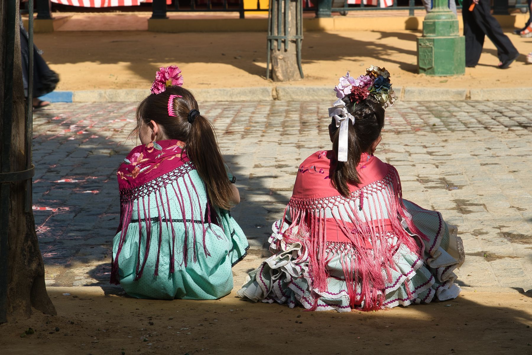 Two girls in flamenco dresses and fringed shawls sitting at the edge of a sandy street at the Seville fair