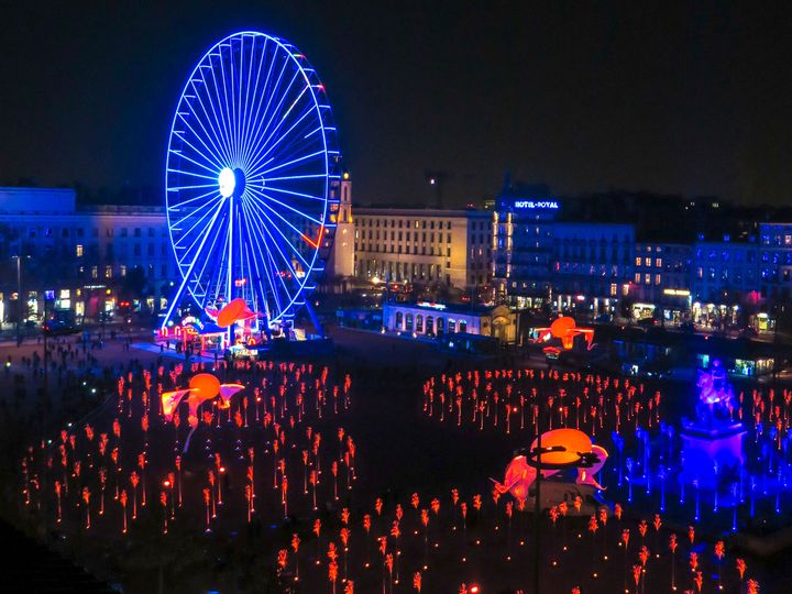 Place Bellecour in Lyon during the Fête des Lumières, filled with red light stems beside a blue-lit Ferris wheel