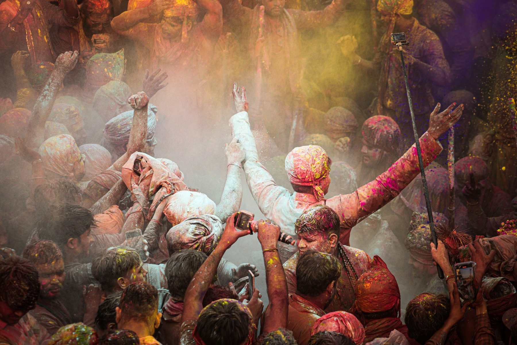 A crowd at Barsana during Holi with arms raised and coloured powder in the air