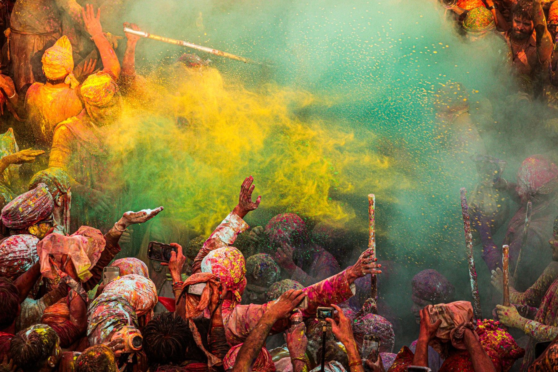 A crowd in Mathura during Holi with a cloud of yellow and green powder bursting over raised arms