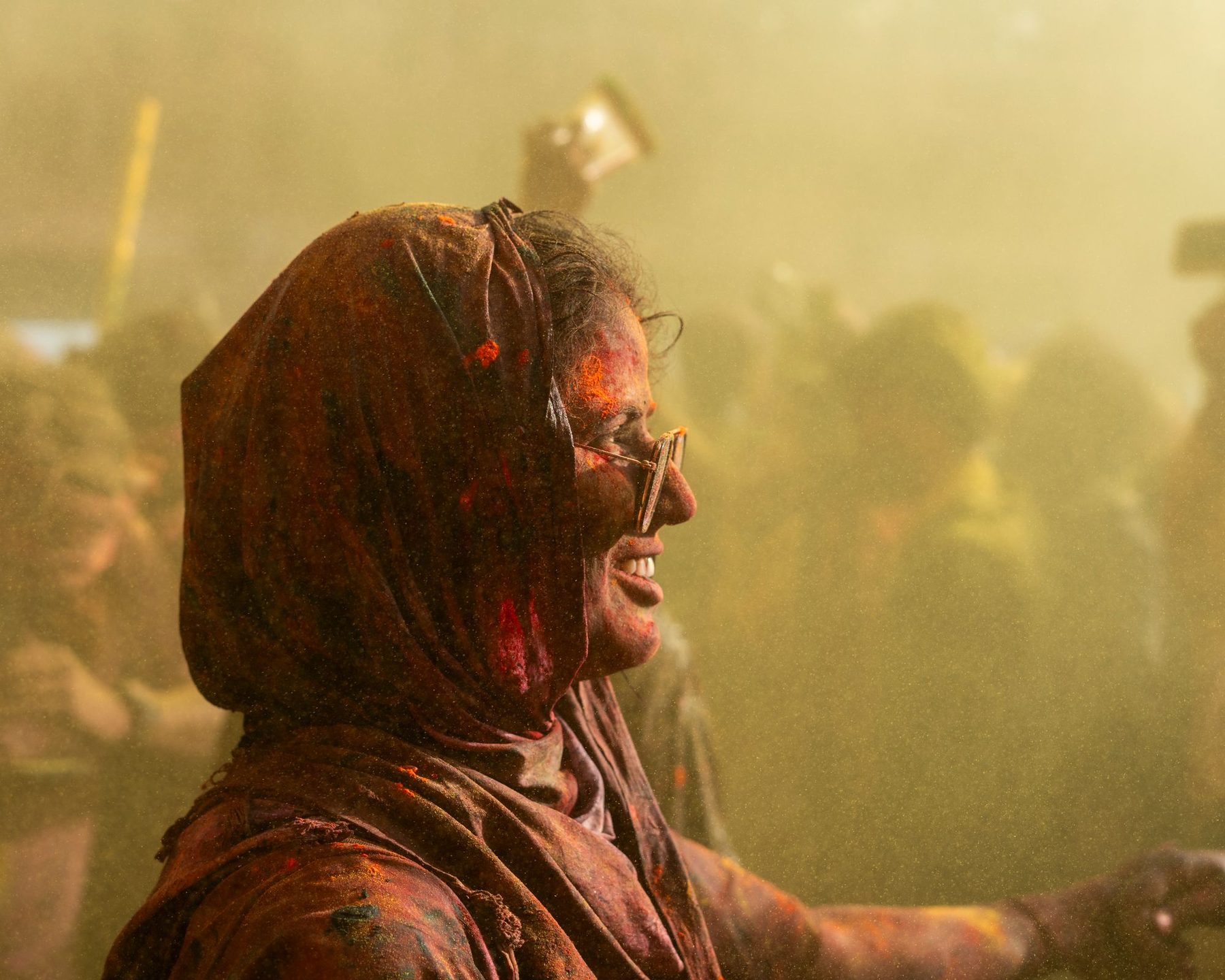 A temple courtyard in Nandgaon during Holi, a woman raised above the crowd on a flagstaff and powder in the air