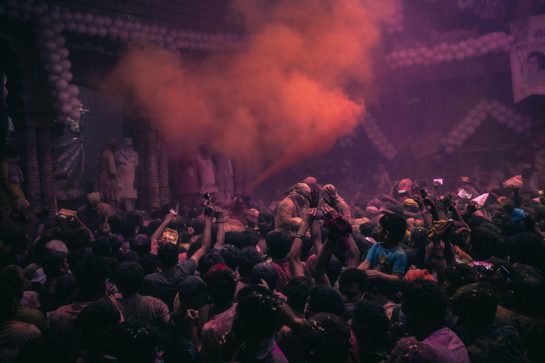 A packed hall in Vrindavan during Holi, hands raised and powder in the air under the lights