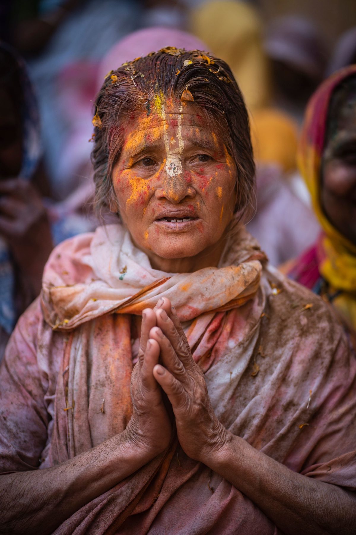 An elderly woman in Vrindavan, her face painted with colour, hands clasped, during Holi
