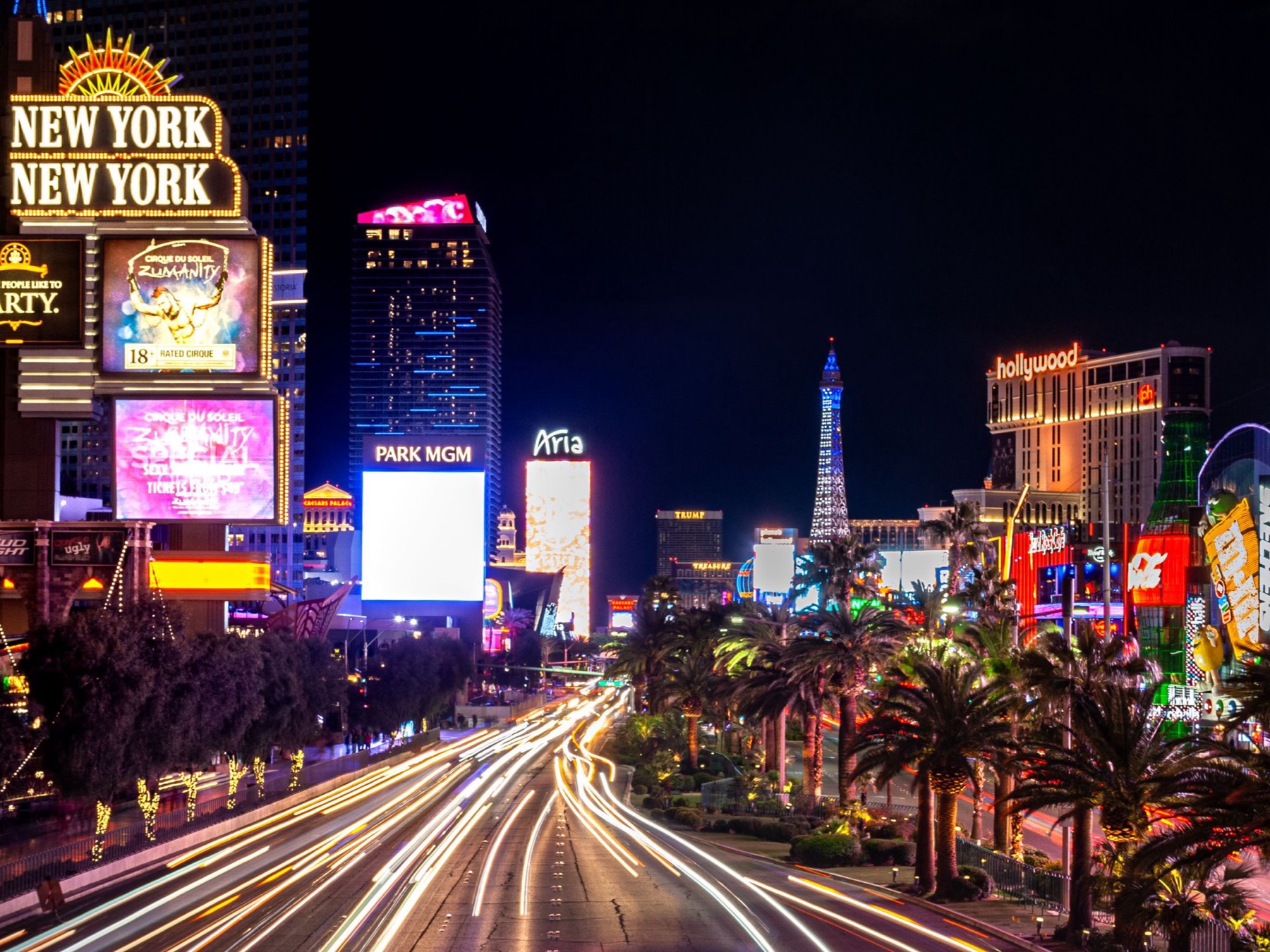 Las Vegas Boulevard at night, looking north from New York-New York past Park MGM, with car light trails either side of the palm-lined median and the Paris hotel's Eiffel Tower replica lit up ahead
