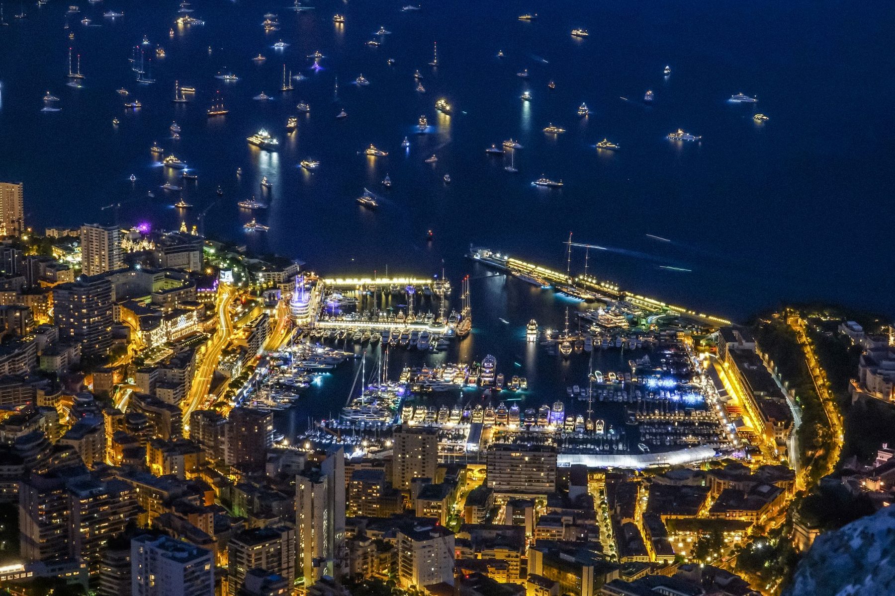 The harbour of Monaco at night from above, the quays lit and dozens of yachts anchored off the port
