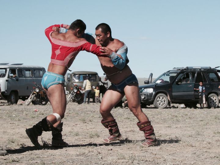 Two wrestlers in traditional boots and jackets grappling on a dusty field at a Mongolian naadam