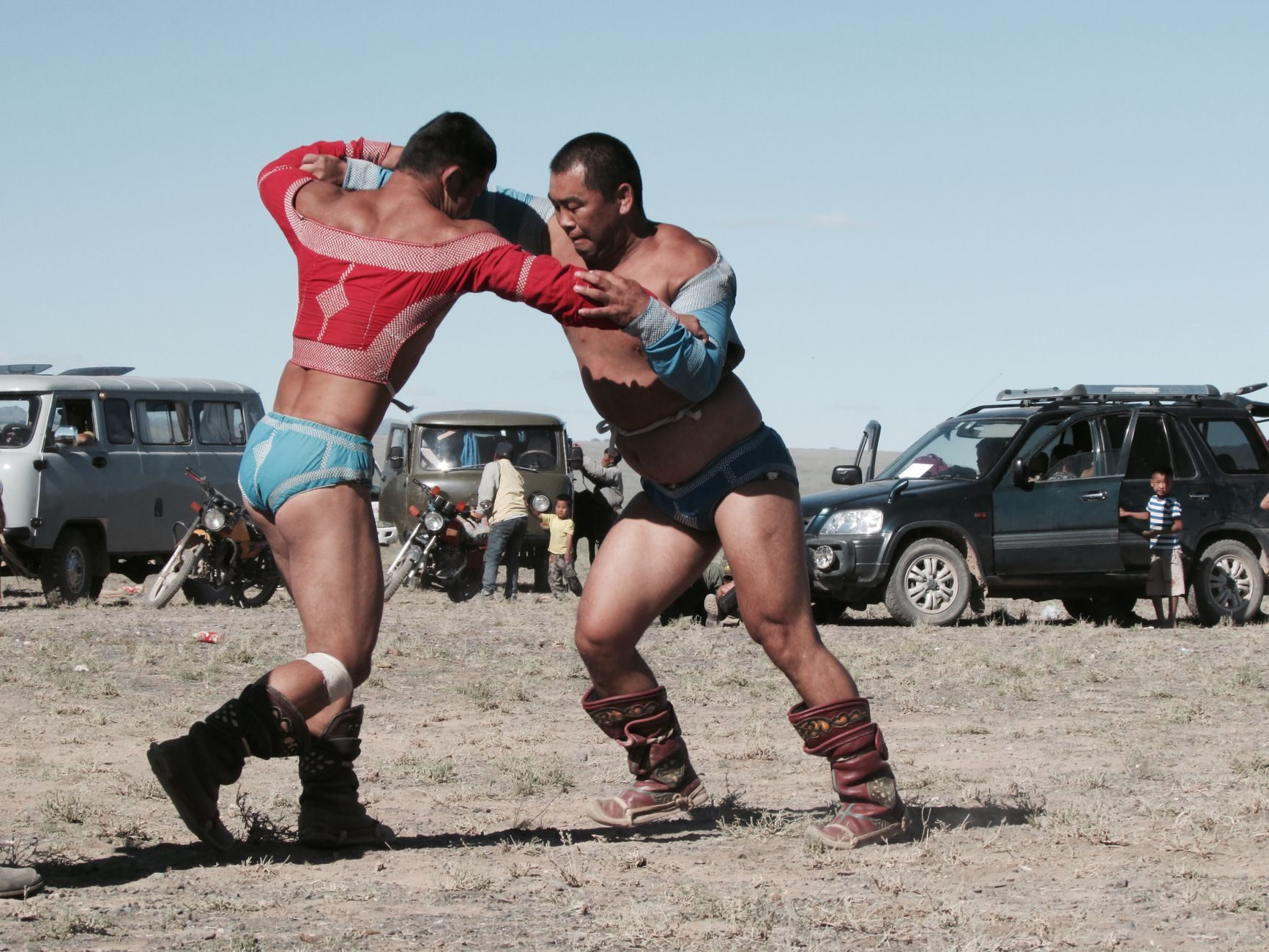Two wrestlers in traditional boots and jackets grappling on a dusty field at a Mongolian naadam