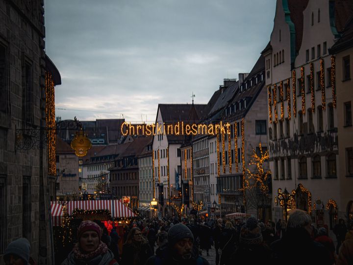 Evening crowds under the lit Christkindlesmarkt sign in Nuremberg's old town, with a red and white striped stall roof on the left