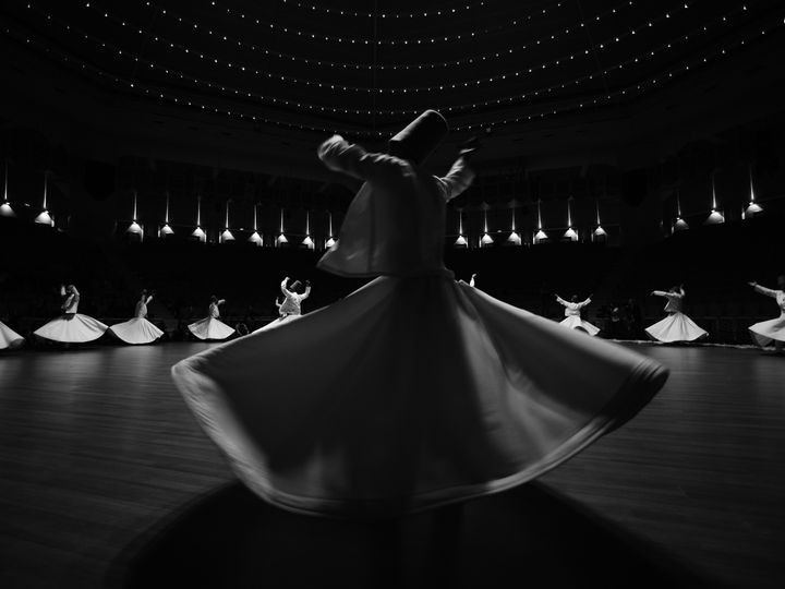 Whirling dervishes in white robes turning under the lit dome of the Sema Hall in Konya, in black and white