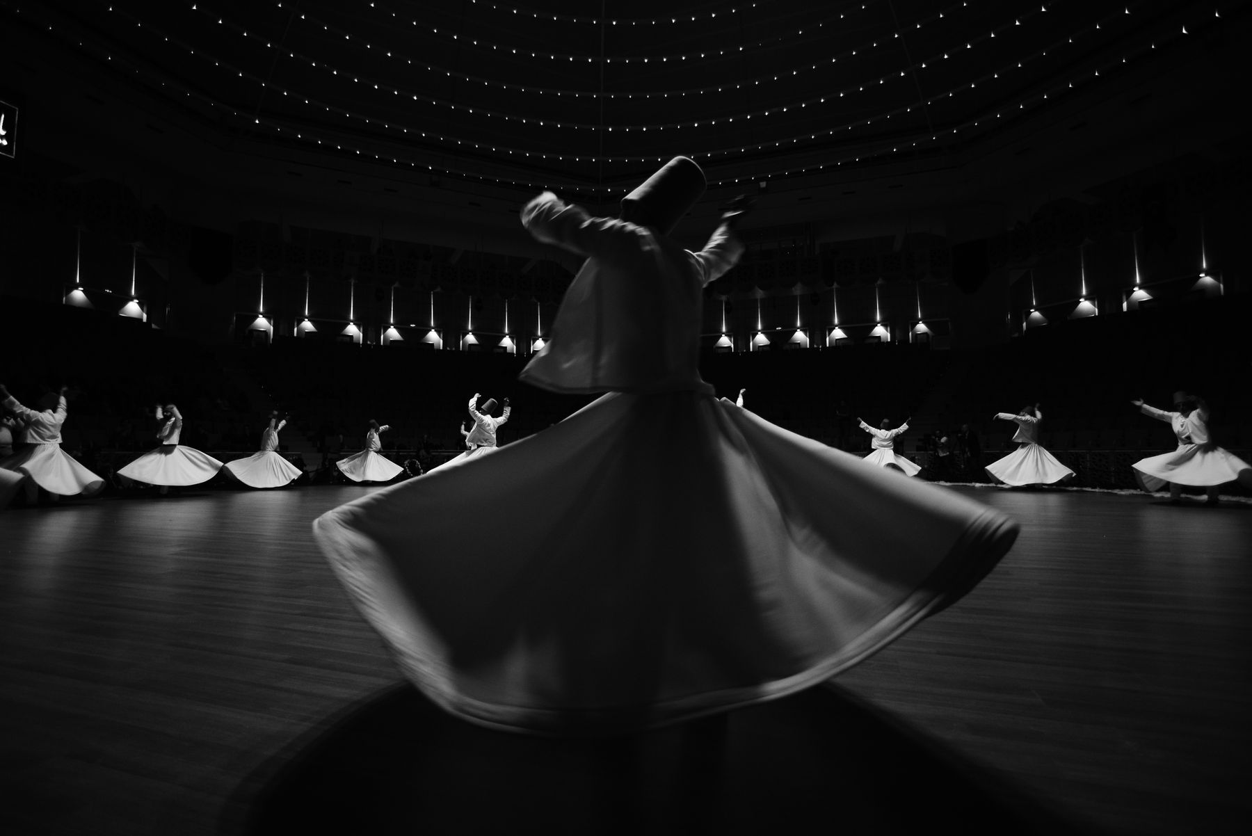 Whirling dervishes in white robes turning under the lit dome of the Sema Hall in Konya, in black and white