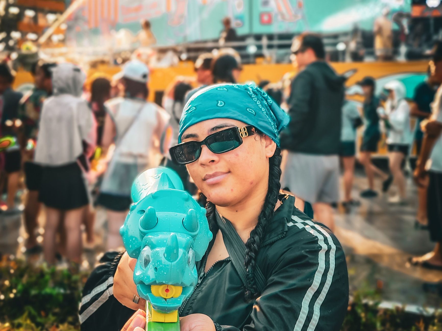 A woman in a bandana and sunglasses holds a water gun towards the camera during Songkran outside the Maya mall in Chiang Mai, with a soaked crowd behind her