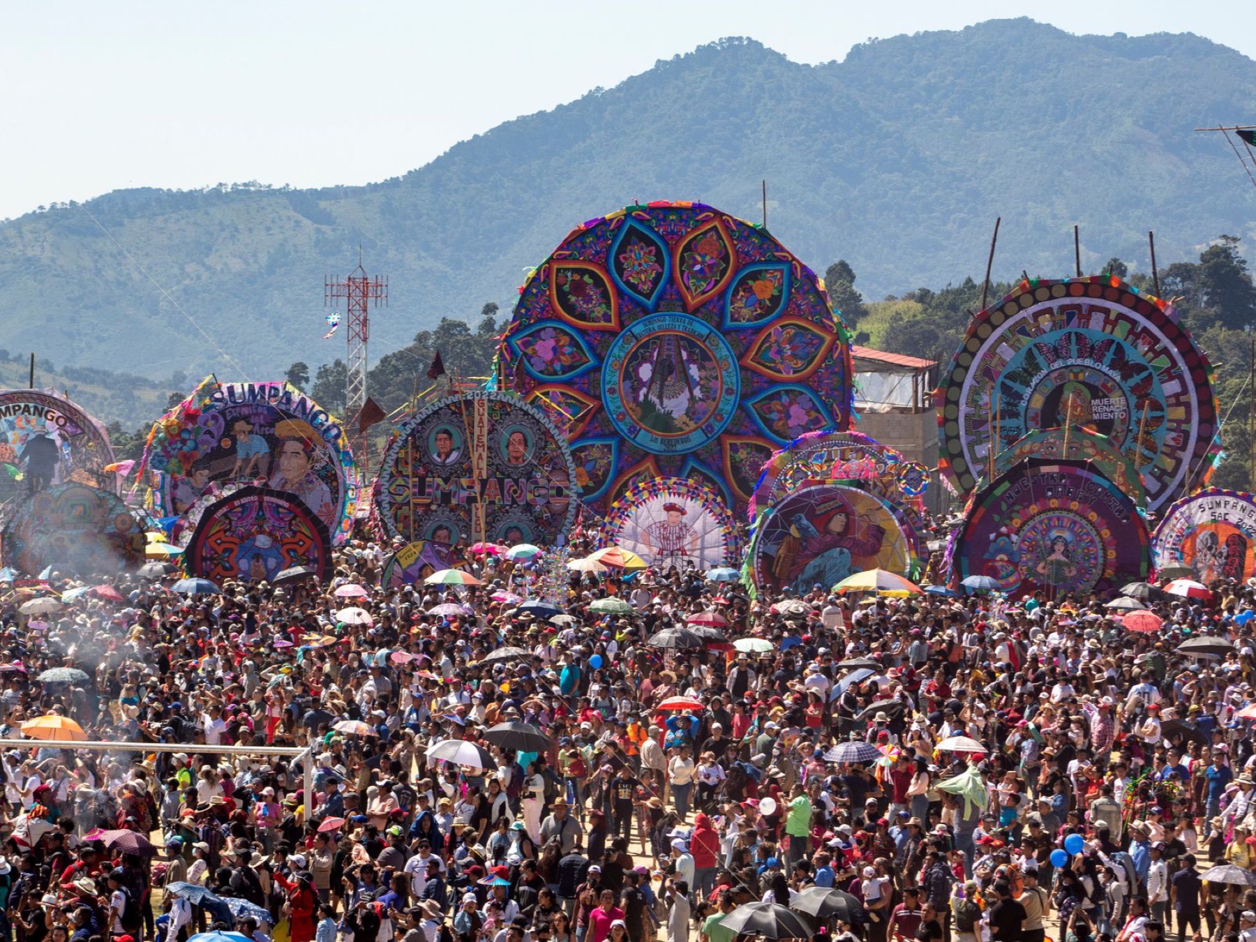 Giant round painted kites standing on poles above a dense crowd on the football field at Sumpango, with green mountains behind