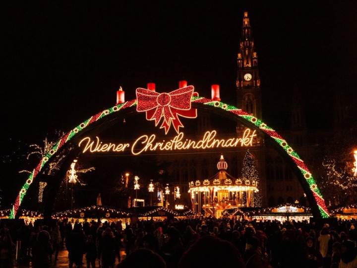 The lit Wiener Christkindlmarkt arch with its red bow above a night crowd on Rathausplatz, a carousel and the Rathaus tower behind
