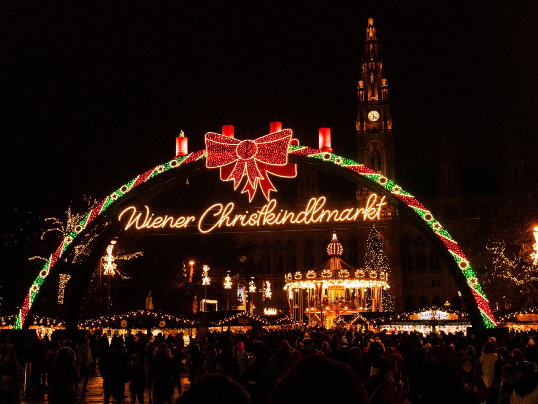 The lit Wiener Christkindlmarkt arch with its red bow above a night crowd on Rathausplatz, a carousel and the Rathaus tower behind