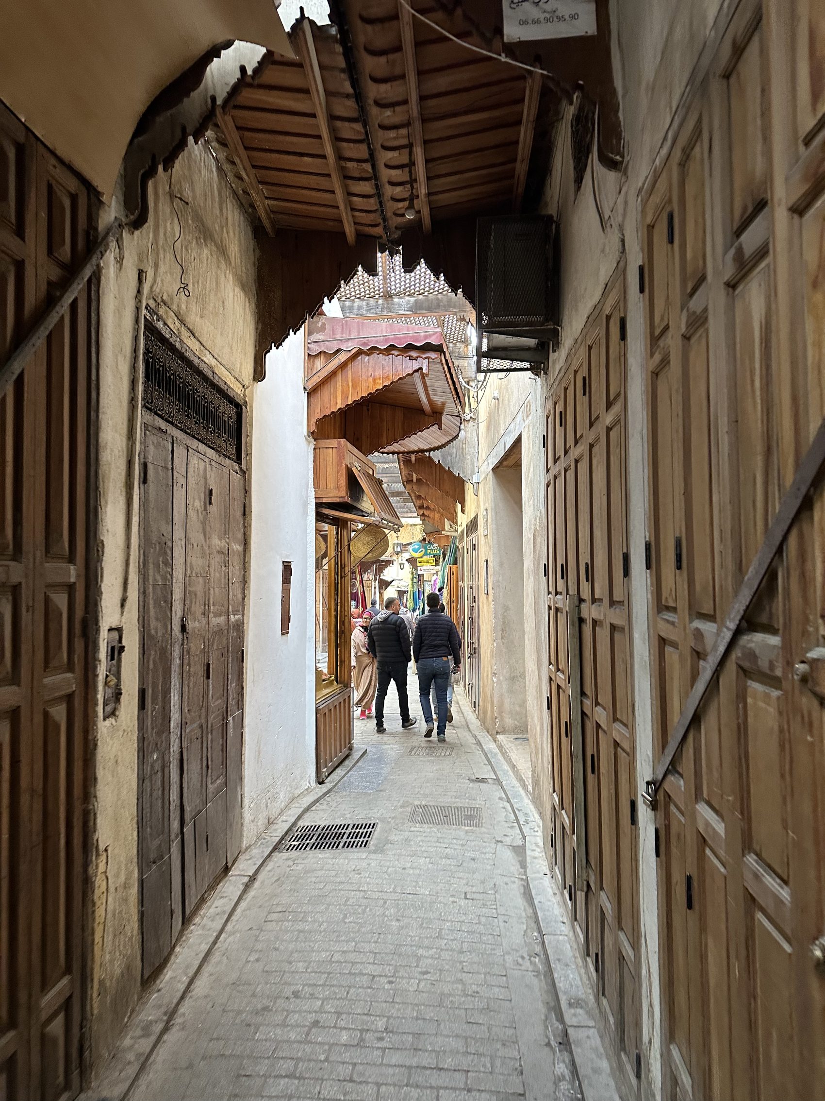 A narrow covered lane in the Fez medina with two figures walking away under a wooden arch
