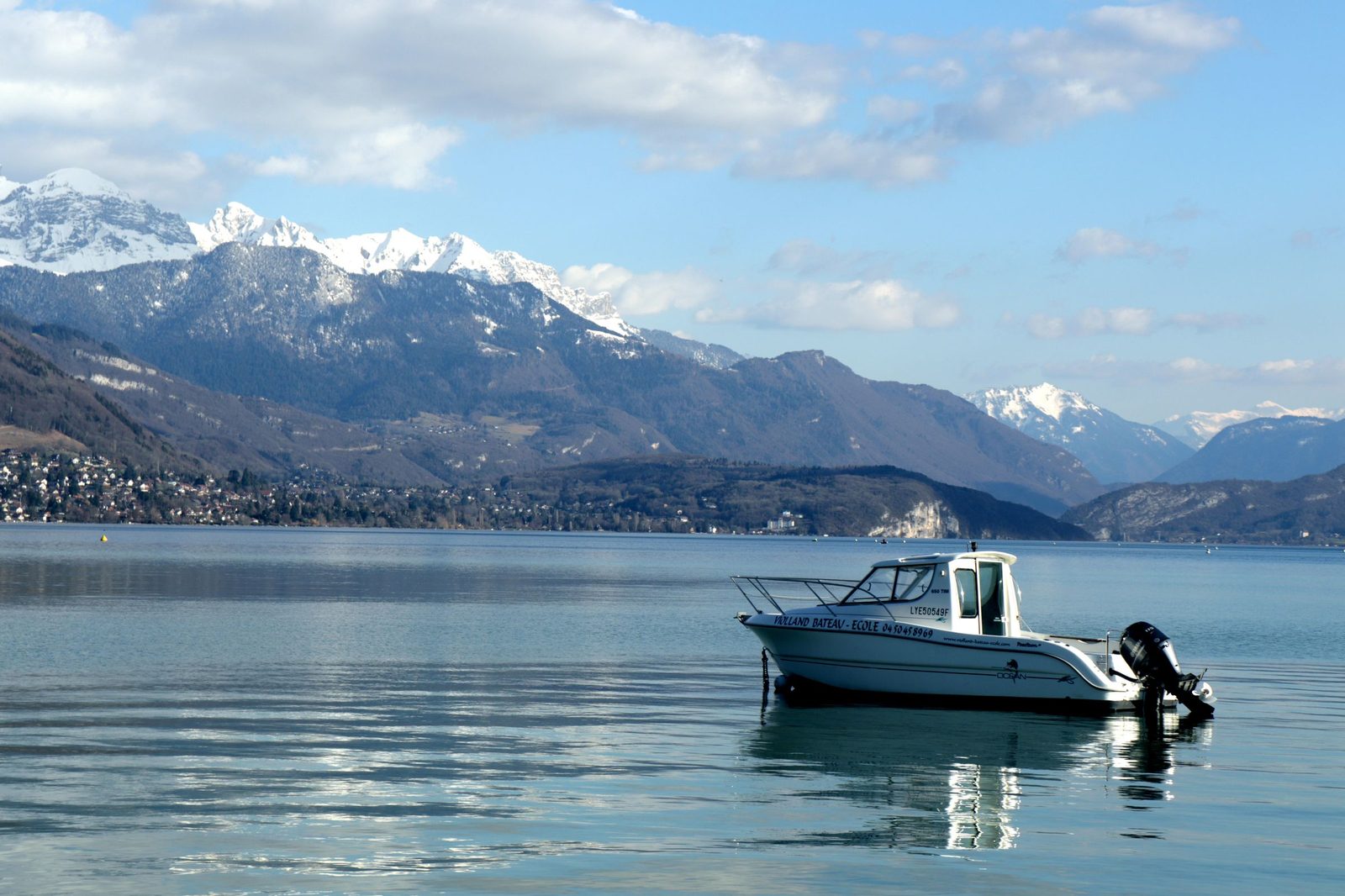 Lake Annecy on a calm day, a small motor boat on the water and snow on the Alps beyond the far shore