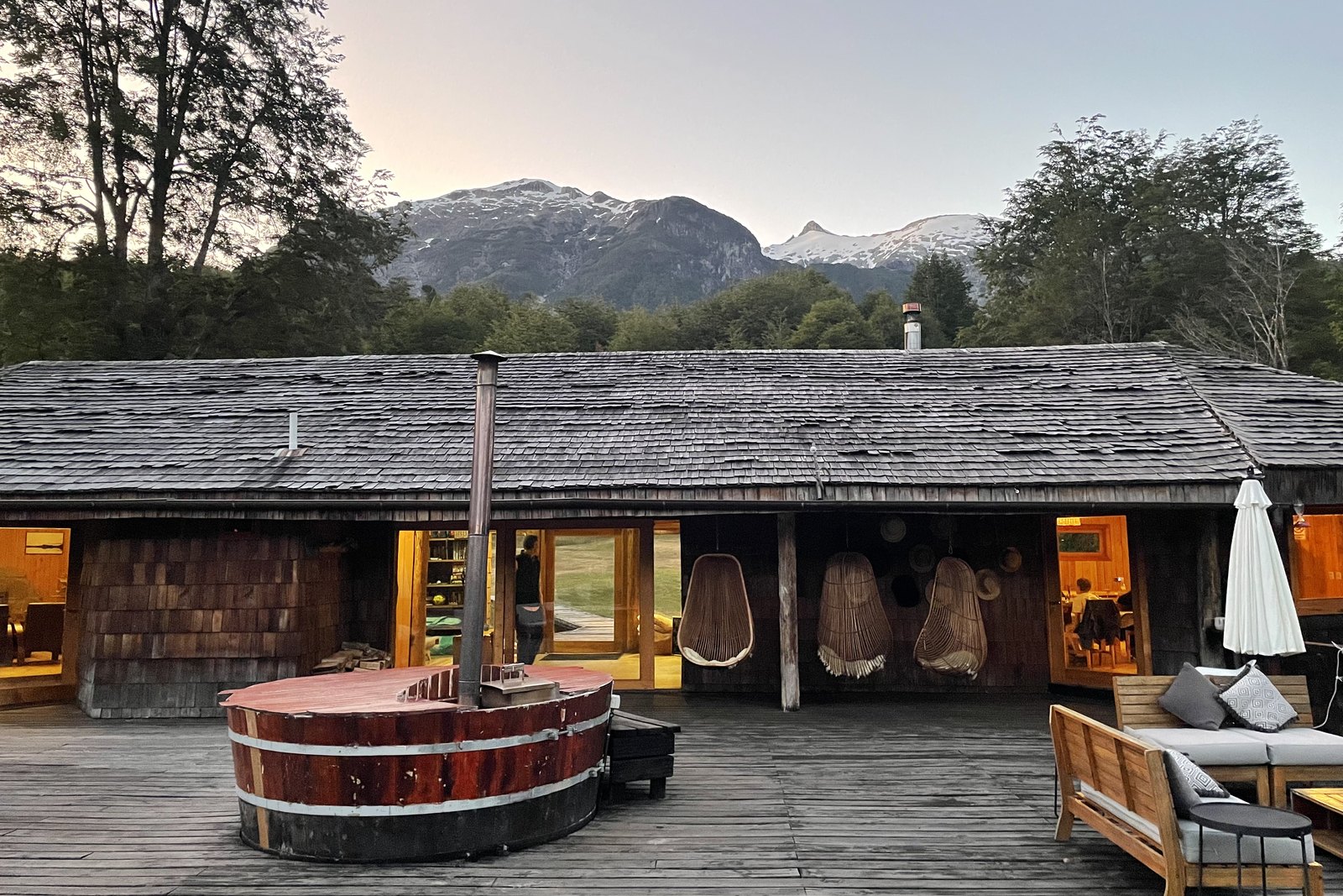 A wood-fired barrel hot tub on the deck of Peuma Lodge at dusk, with snow-capped mountains behind