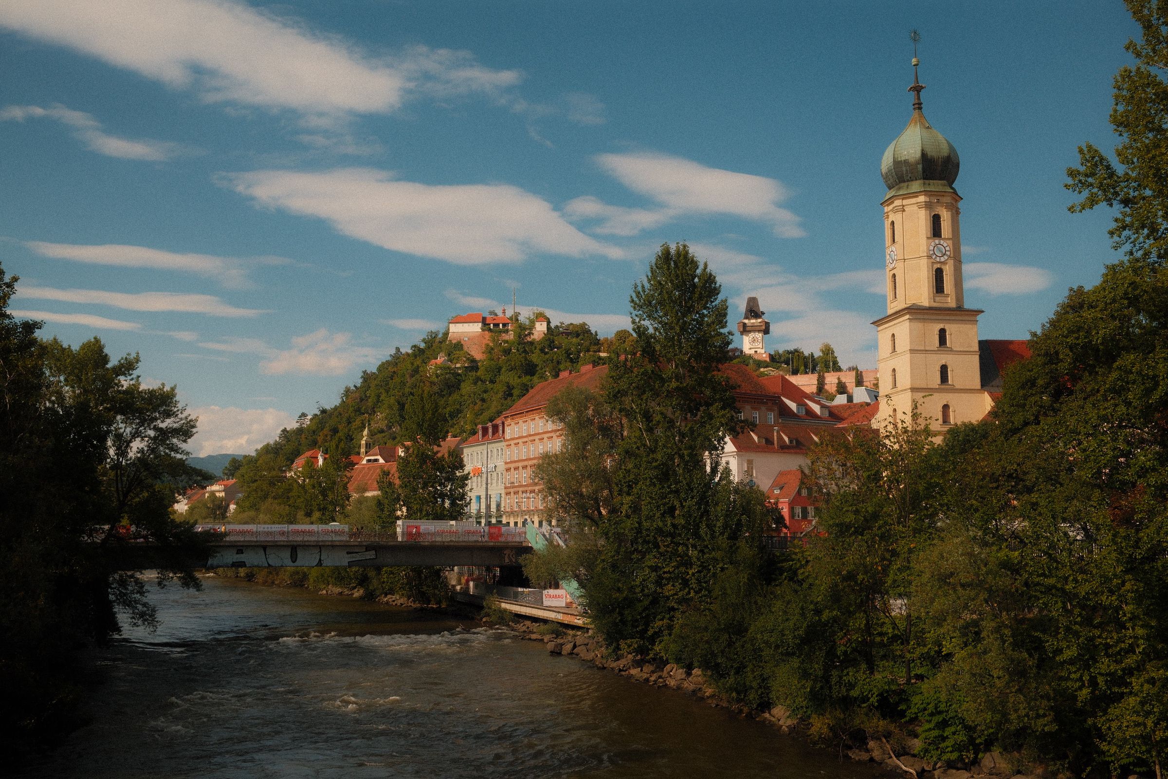 The Mur river in Graz with a road bridge, the wooded Schlossberg behind it with the small Uhrturm clock tower near the top, and a church tower with a green onion dome on the right