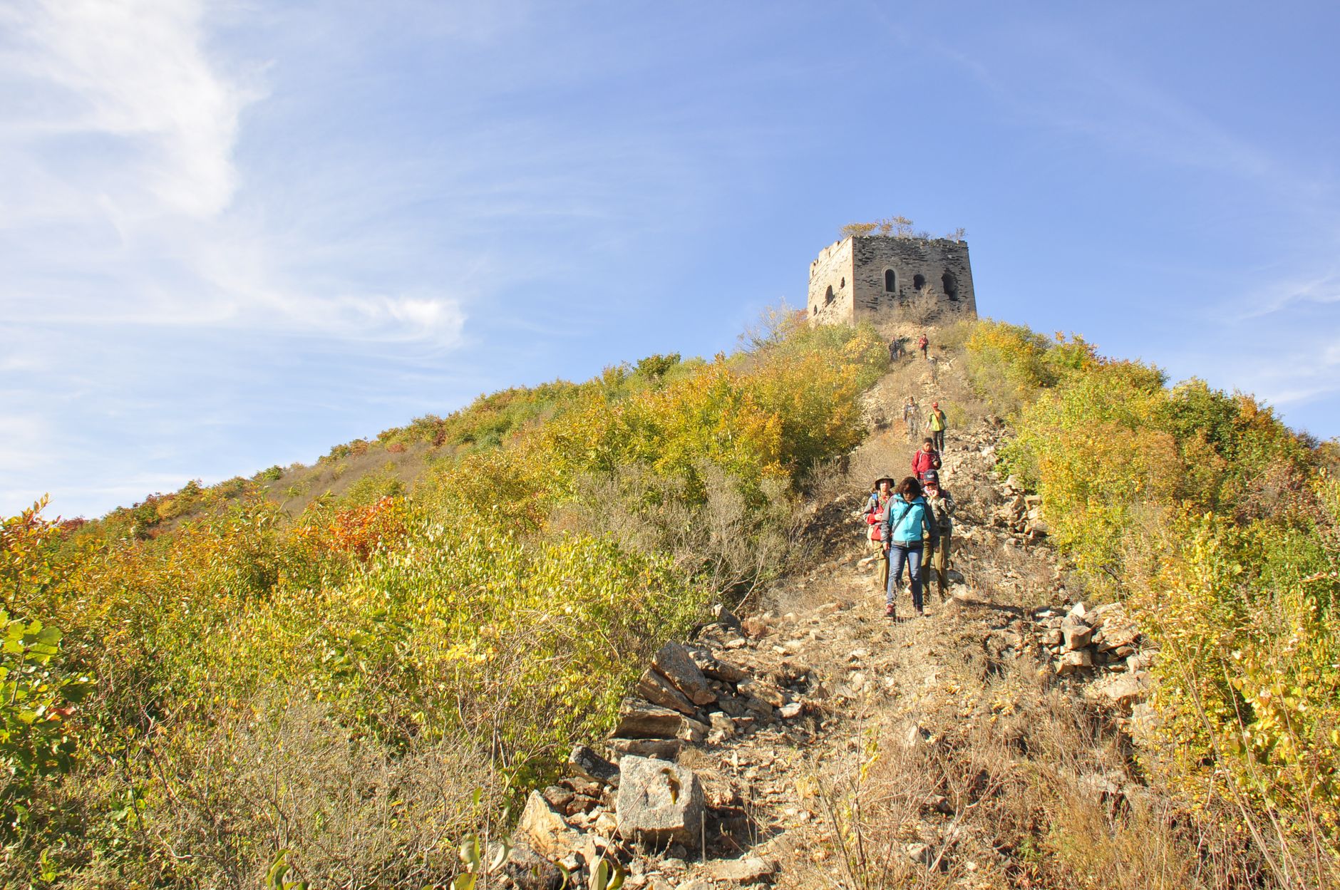 Hikers climbing a rough trail toward a ruined stone tower on a hilltop in autumn