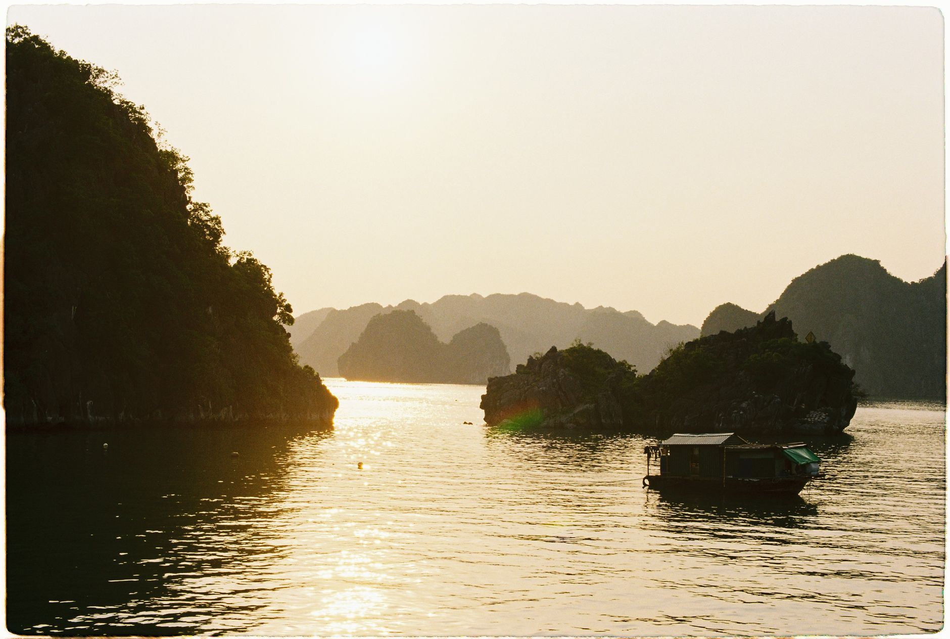 Low sun behind limestone islands in Bai Tu Long Bay with a single small boat on still water