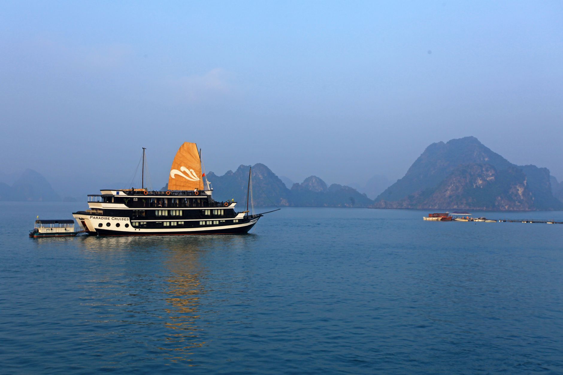 A cruise boat with a tan sail crossing open water between limestone karst islands in Ha Long Bay