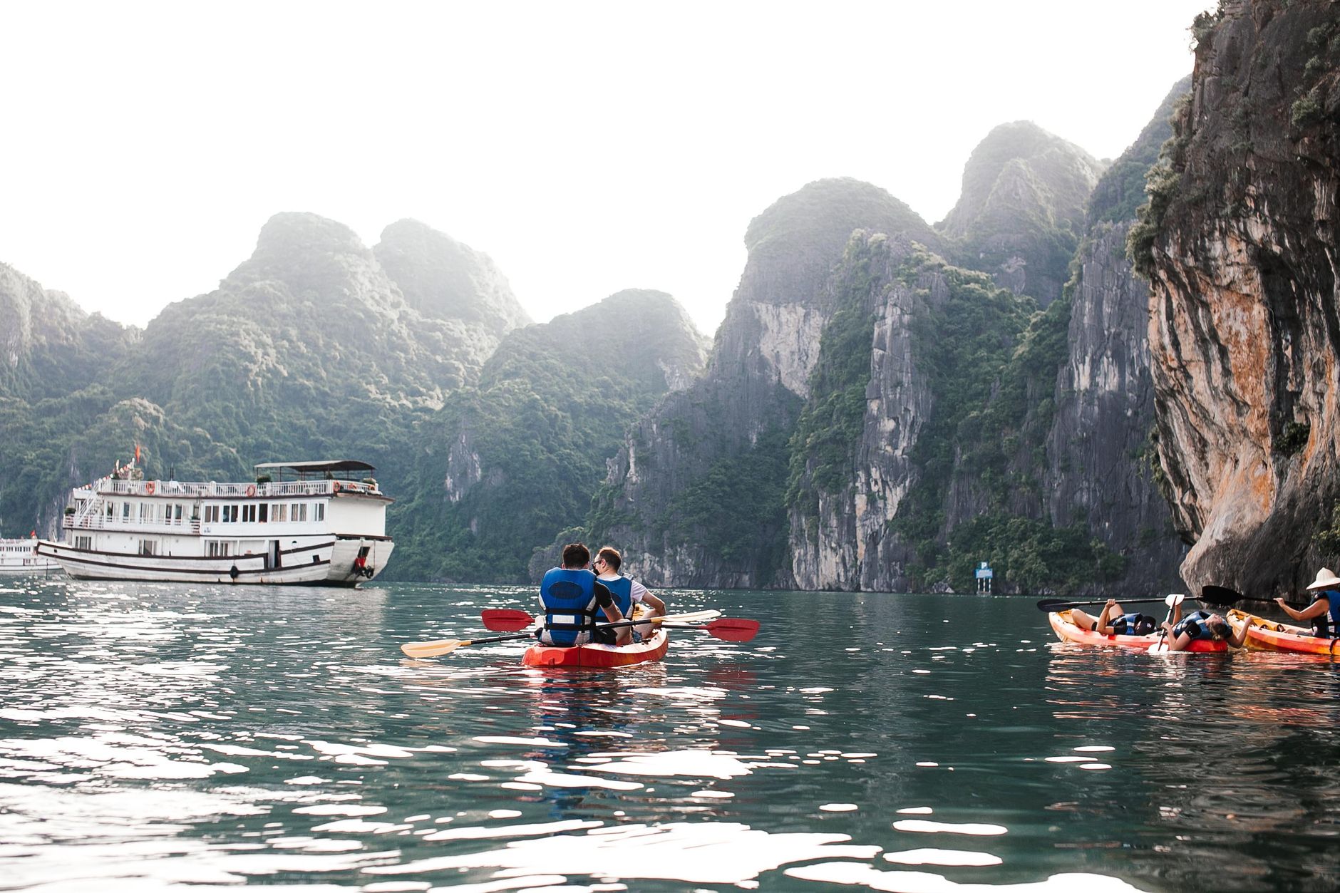 Kayakers paddling beneath limestone cliffs in Ha Long Bay with a white cruise boat anchored behind them