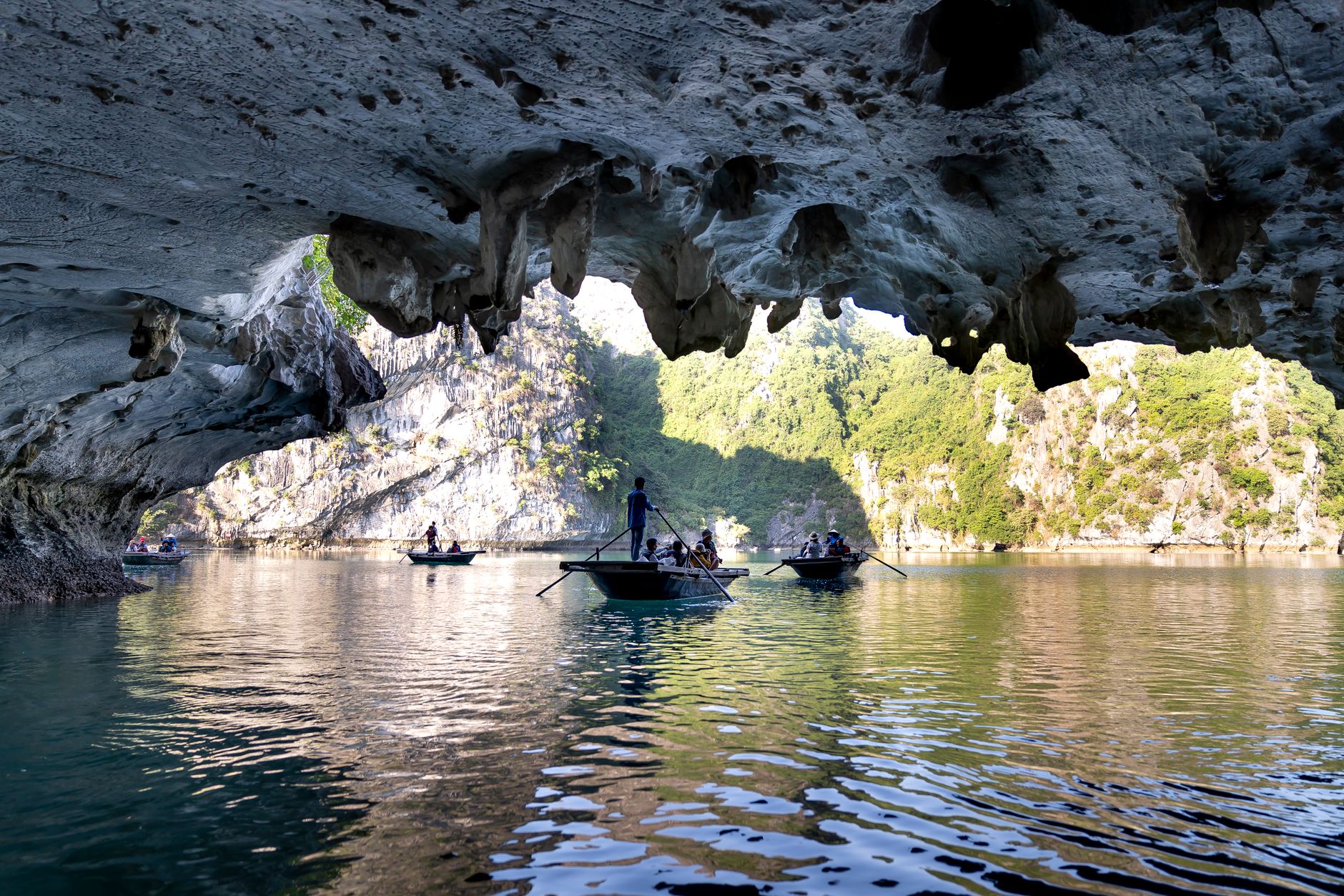 Small wooden boats passing beneath a low limestone arch into a hidden lagoon in Ha Long Bay
