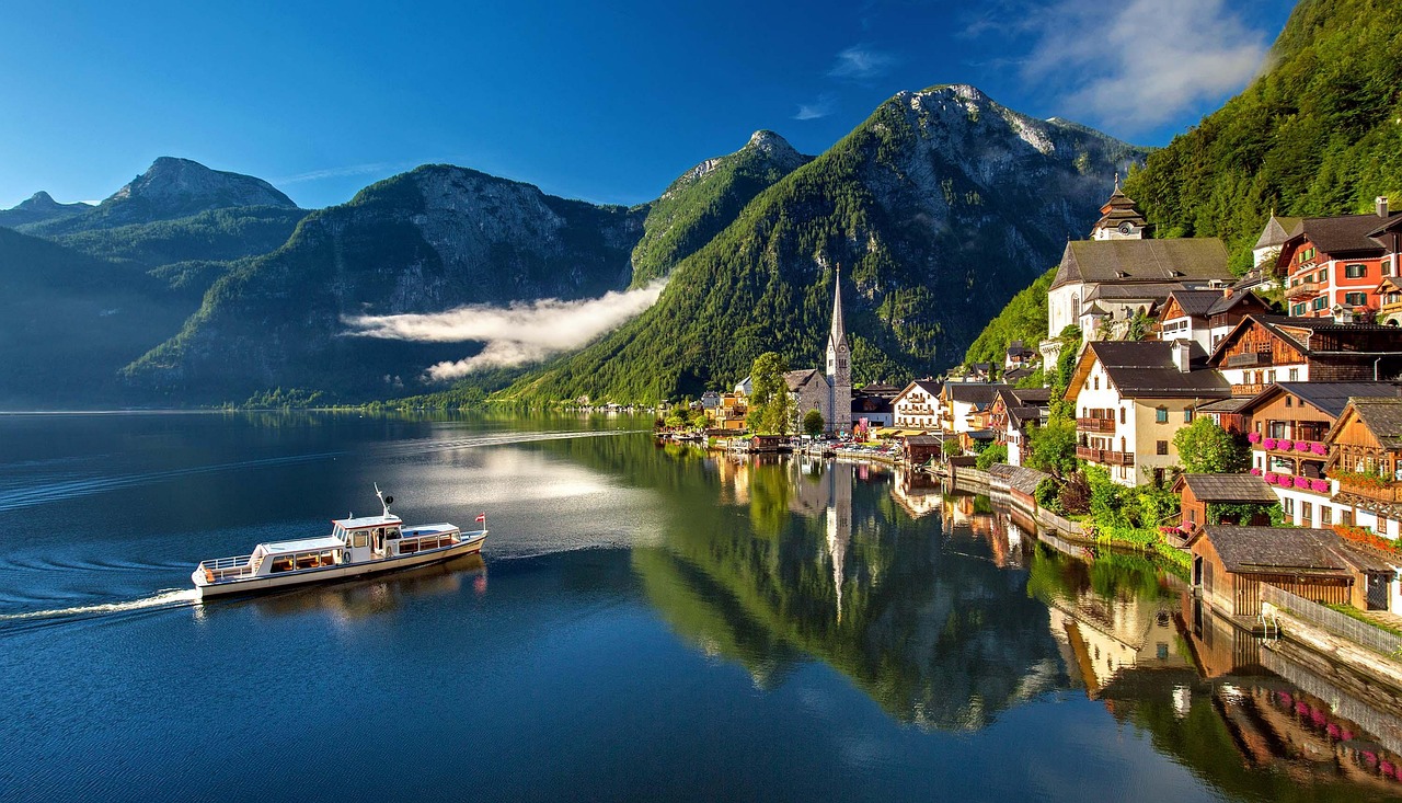 Hallstatt reflected in the flat water of the Hallstattersee with a small passenger ferry crossing and cloud caught on the wooded mountainside behind