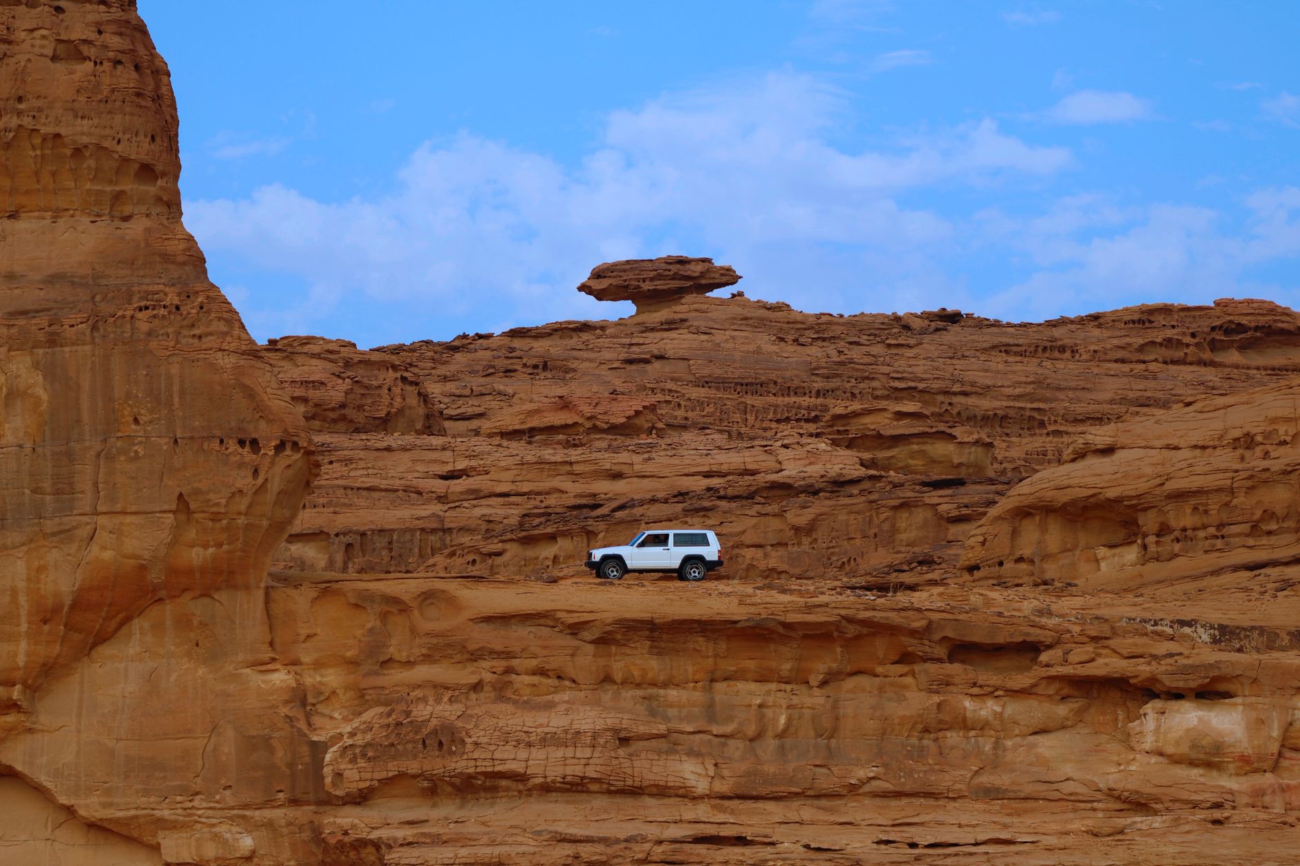 A white vehicle on a ledge beneath layered sandstone cliffs in the AlUla valley, Saudi Arabia