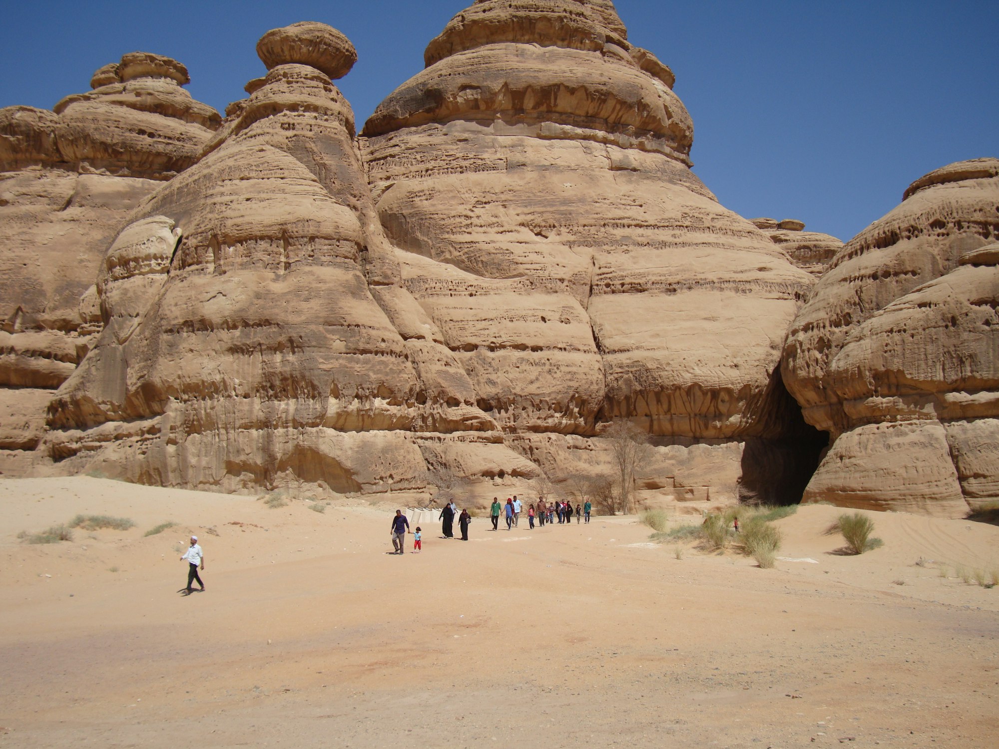 A group of visitors walking across open sand toward large weathered sandstone outcrops at Hegra, Saudi Arabia