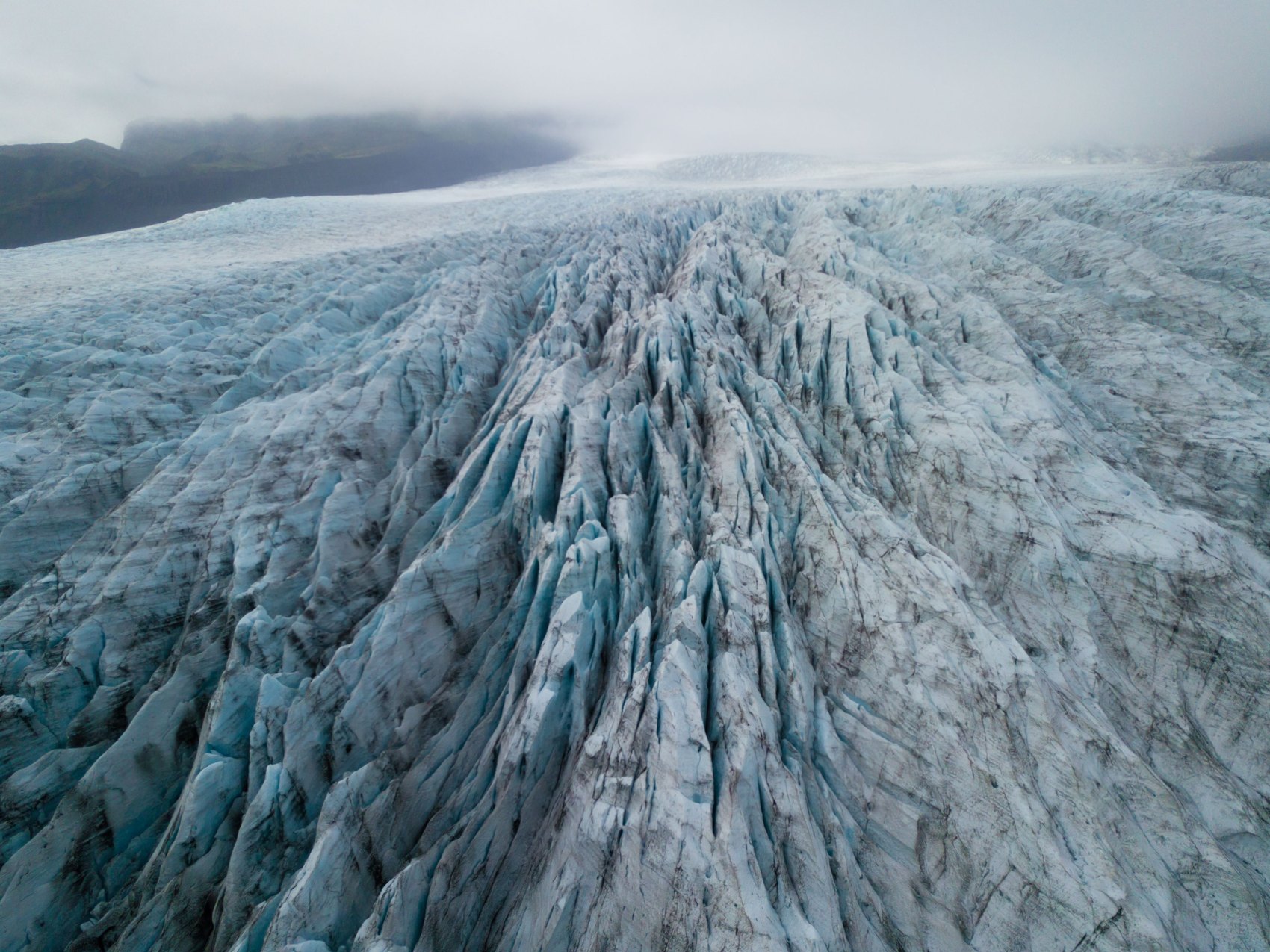 A crevassed glacier surface of blue and grey ice streaked with dark ash, running away towards low cloud