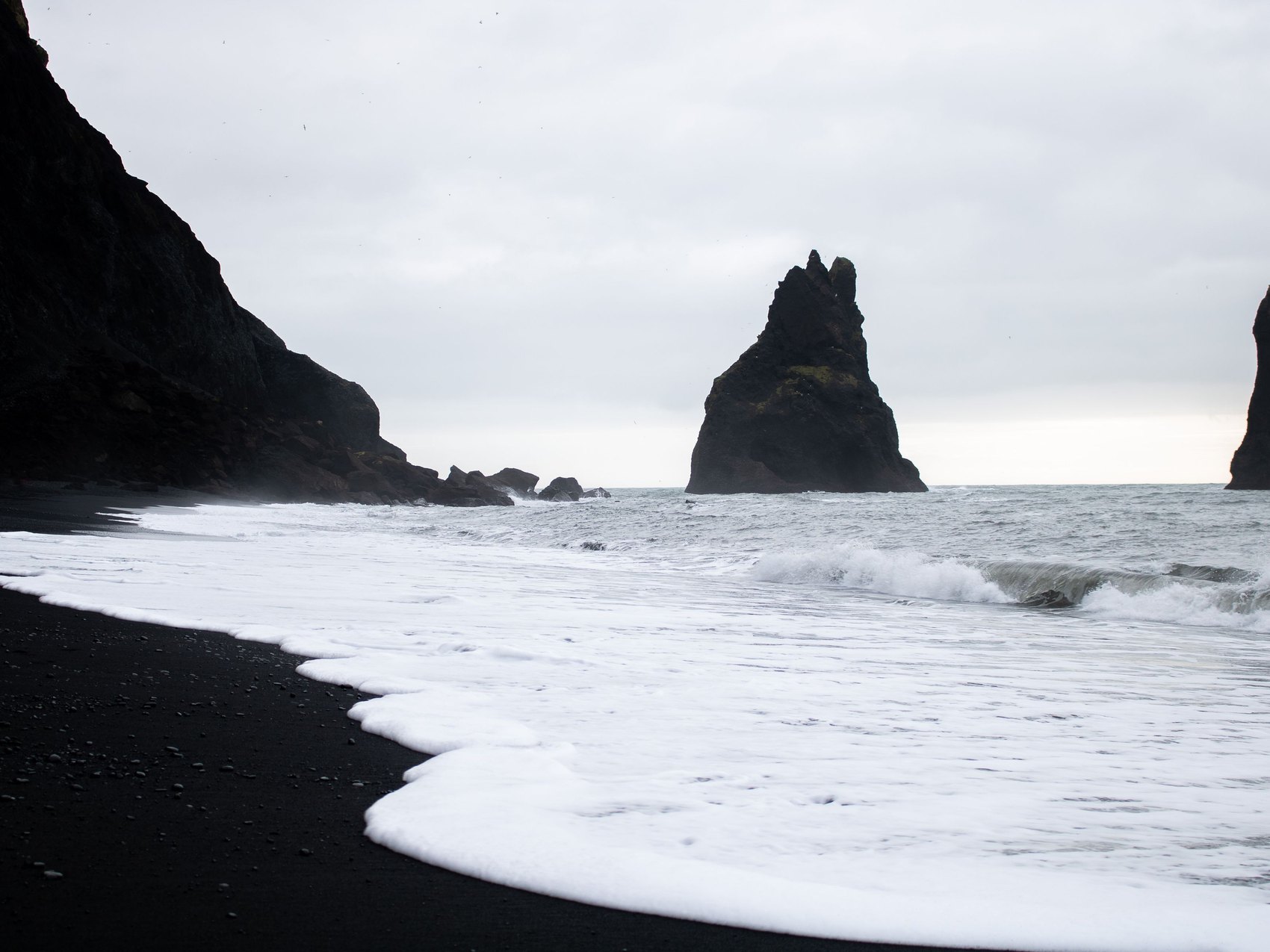 Black sand and white surf running up a beach, with two dark sea stacks standing offshore under a pale grey sky
