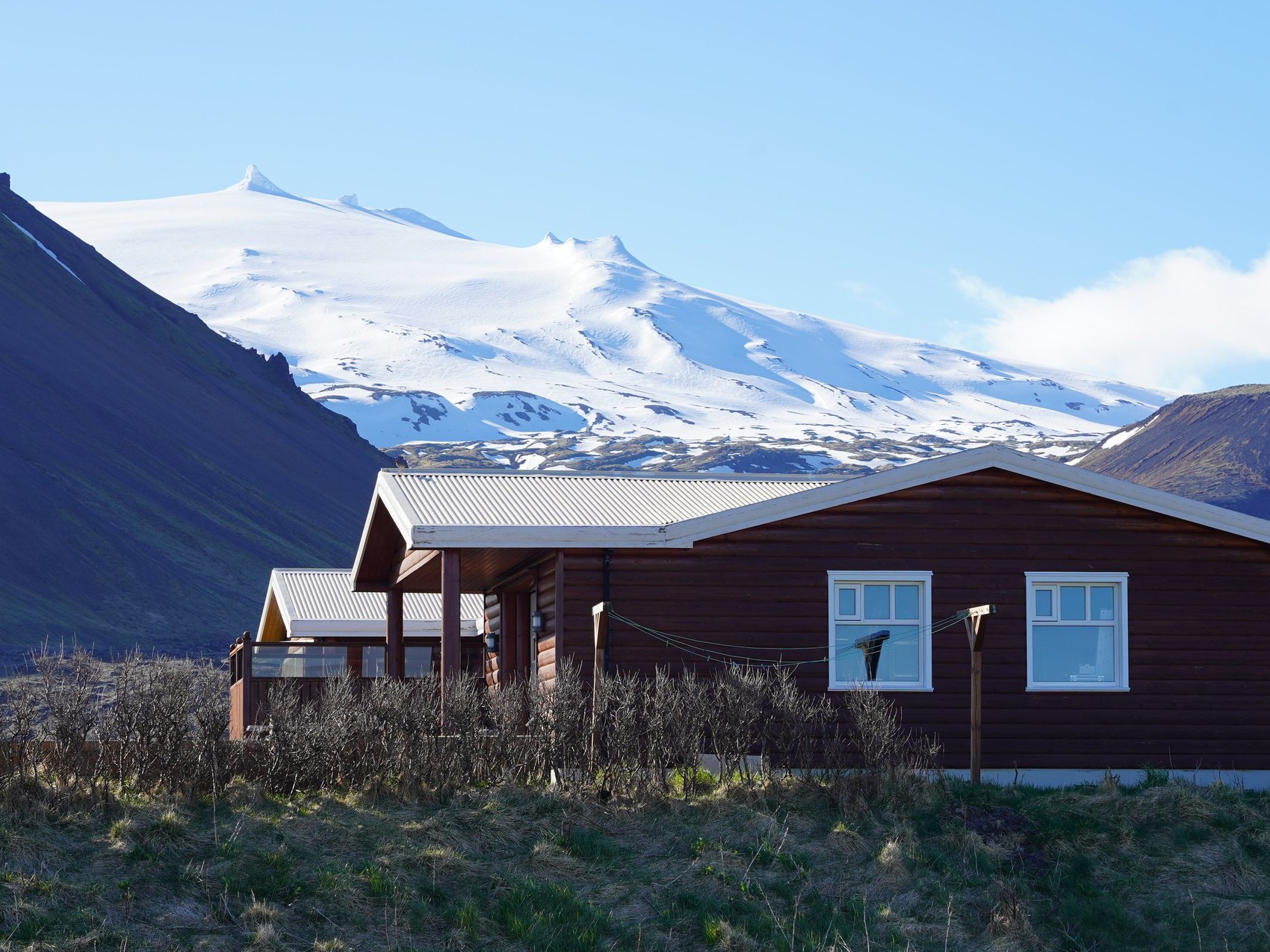 A dark timber lodge with white window frames standing alone in rough grass below a broad snow covered ice cap under a clear sky