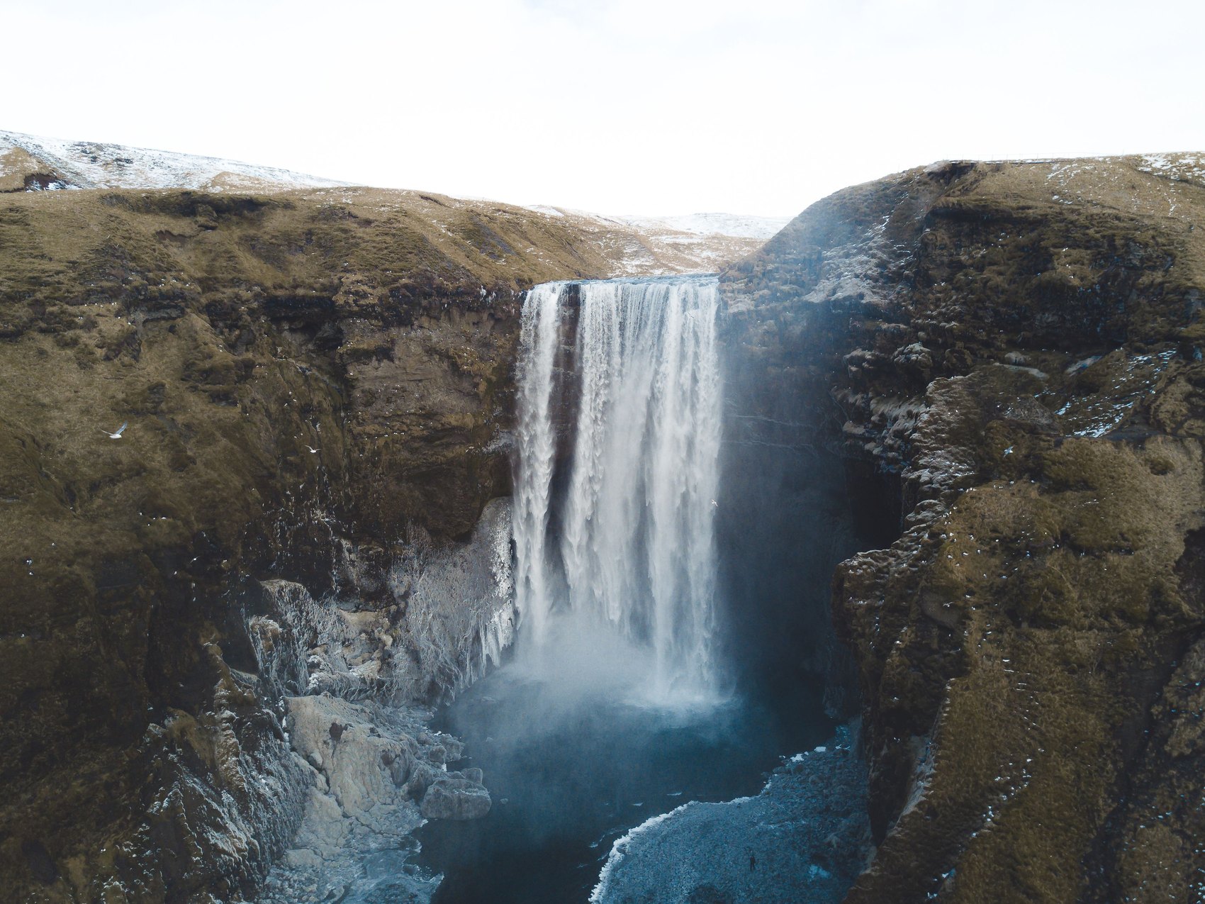An aerial view of a wide waterfall dropping over a dark cliff into a pool, with flat snow streaked moorland running away on both sides