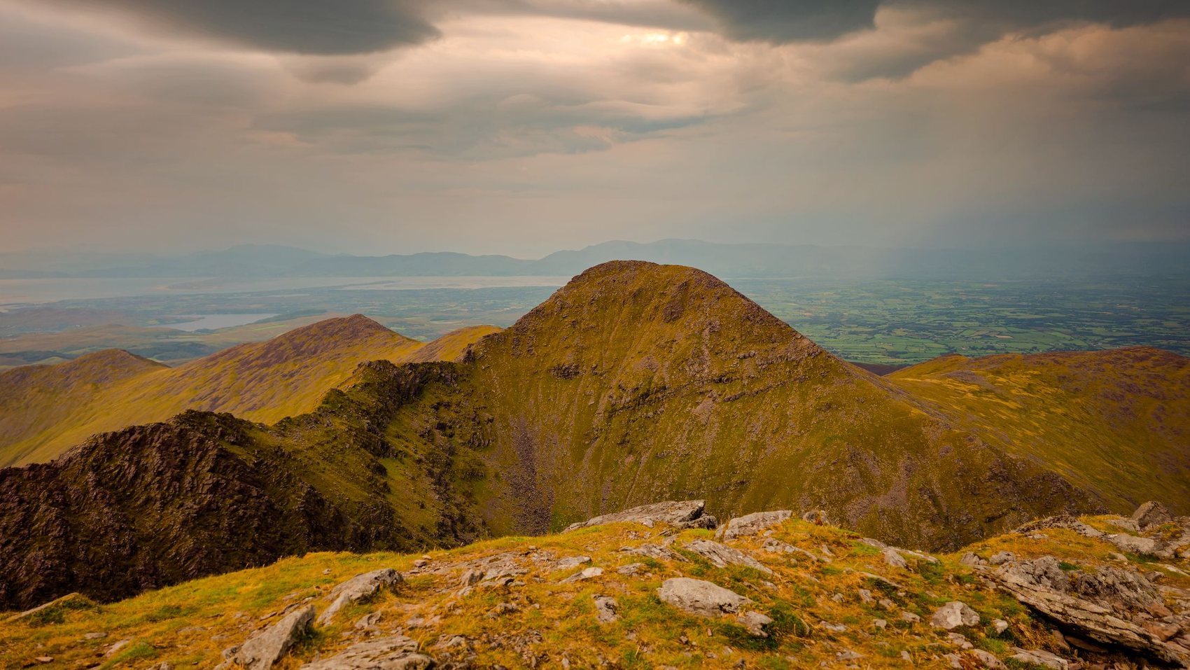 A grassed rocky ridge of the MacGillycuddy's Reeks seen from high on the range, with storm light breaking over the plain and the sea far behind