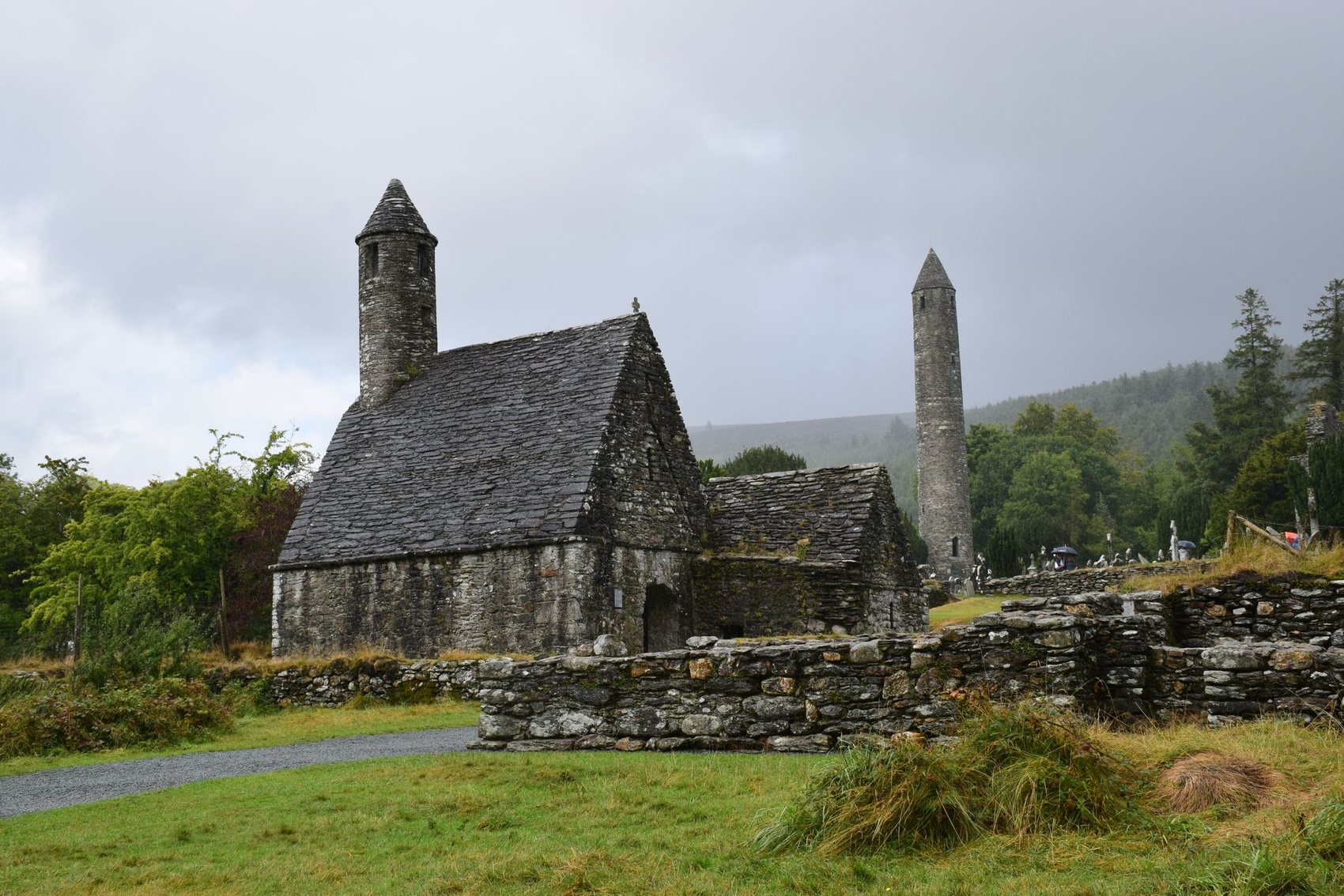 A steep stone-roofed church and a tall round tower standing in the wooded valley at Glendalough under grey cloud, behind a low drystone enclosure wall