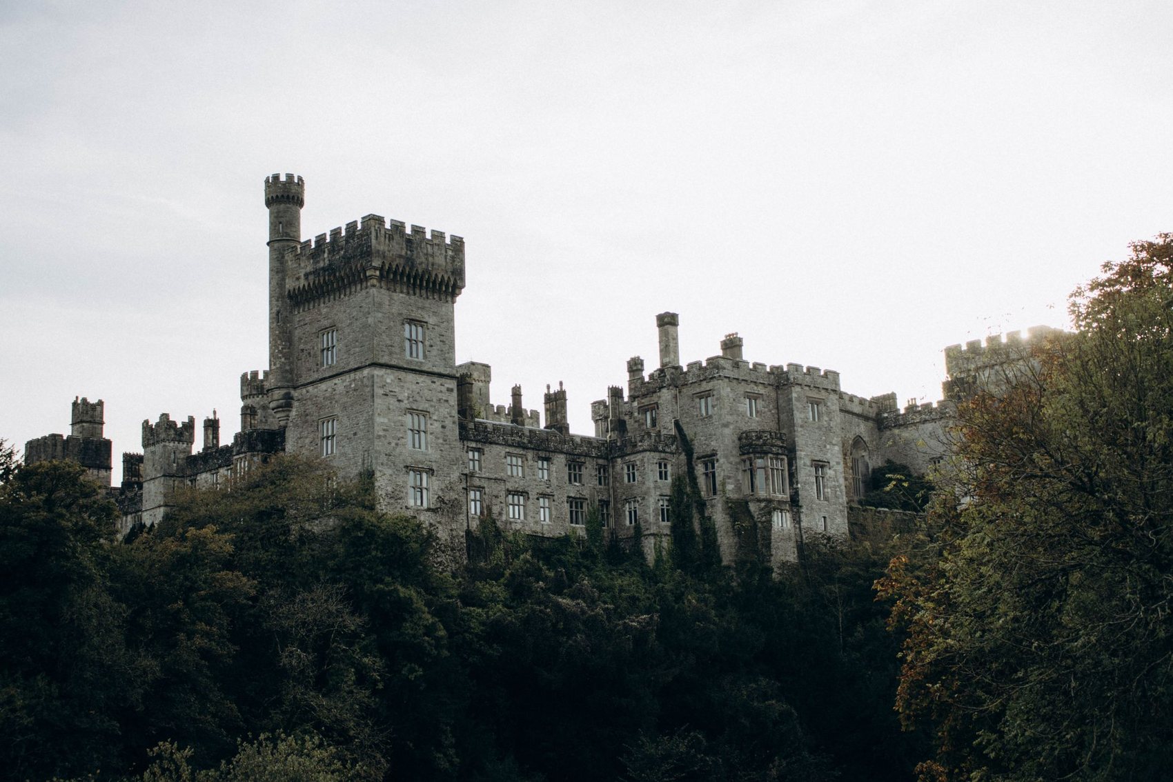 The battlemented towers and turrets of Lismore Castle rising out of dense woodland, under a pale overcast sky
