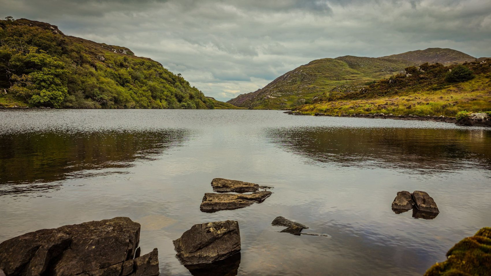 A still lake under heavy grey cloud in the Killarney hills, wooded on the near shore and bare rock and heather on the far one, with dark stones in the foreground