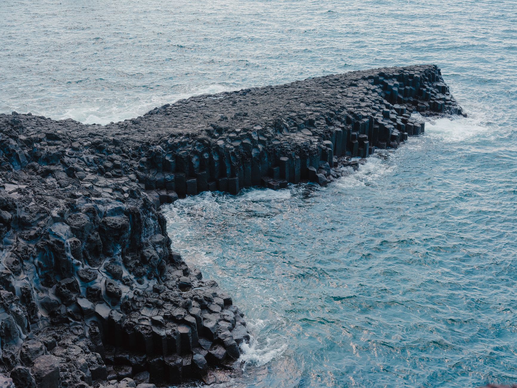 Columnar basalt forming a low dark promontory into green-blue sea, seen from above