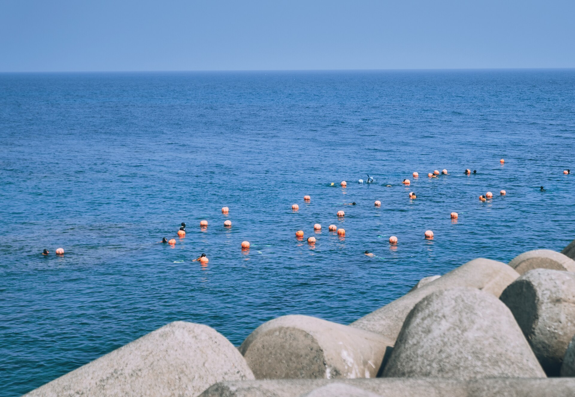 Dozens of orange floats spread across flat blue water with black-hooded divers between them, concrete tetrapods in the foreground