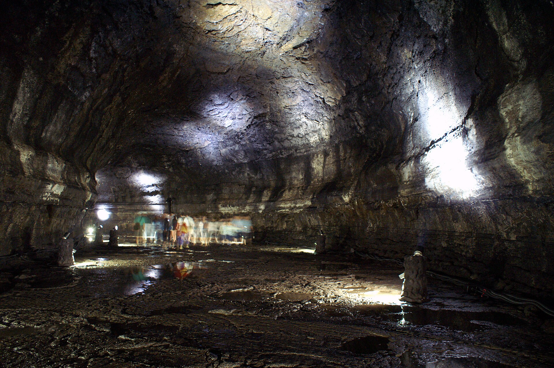 A broad high wet-floored lava tube passage with a string of visitor lights receding into the dark