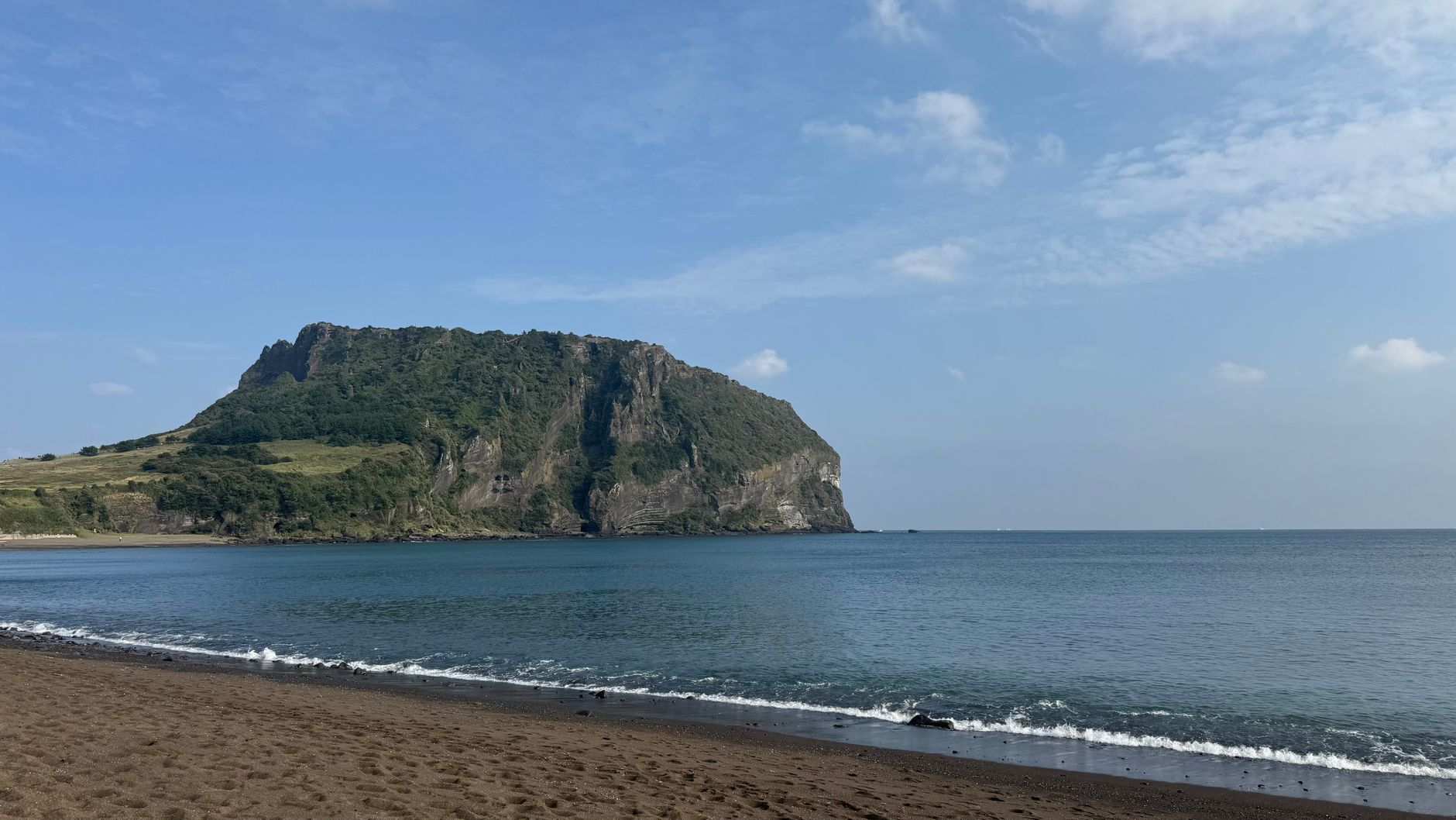 The Seongsan Ilchulbong tuff cone seen across the water from a black-sand beach in flat morning light
