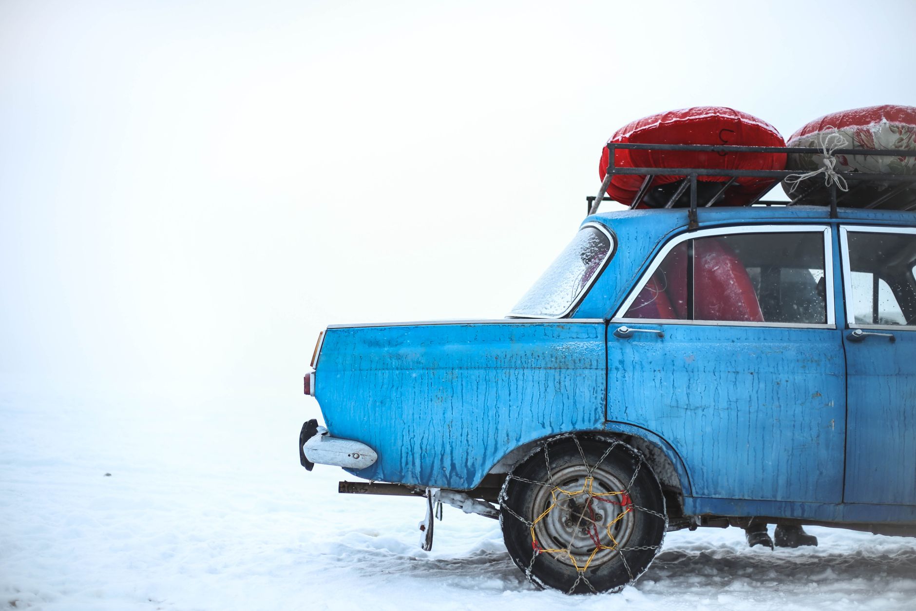 A vintage blue car with red luggage strapped to its roof, parked in deep snow