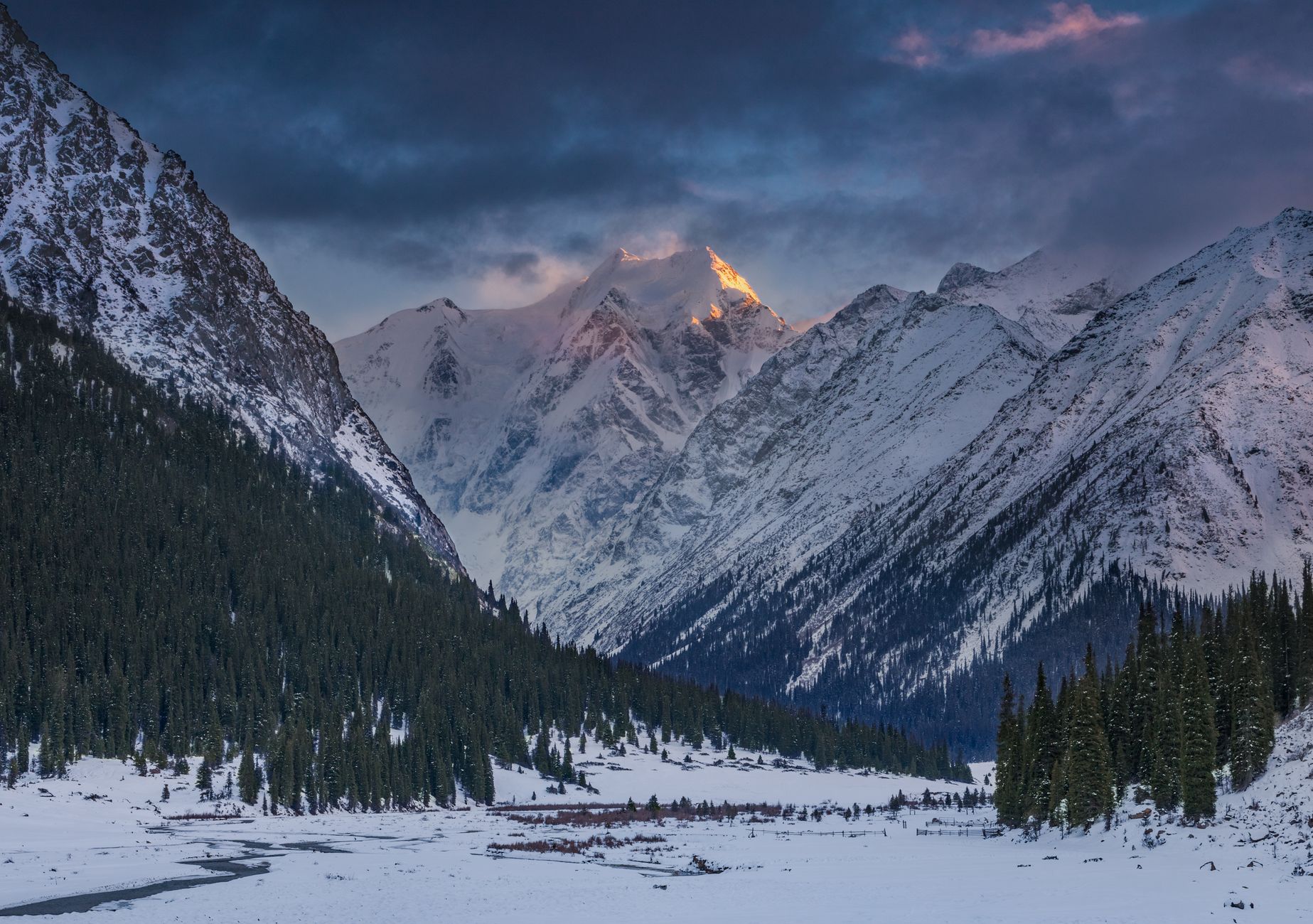 A dark mountain gorge with snowy peaks lit gold by sunrise above a frozen valley floor
